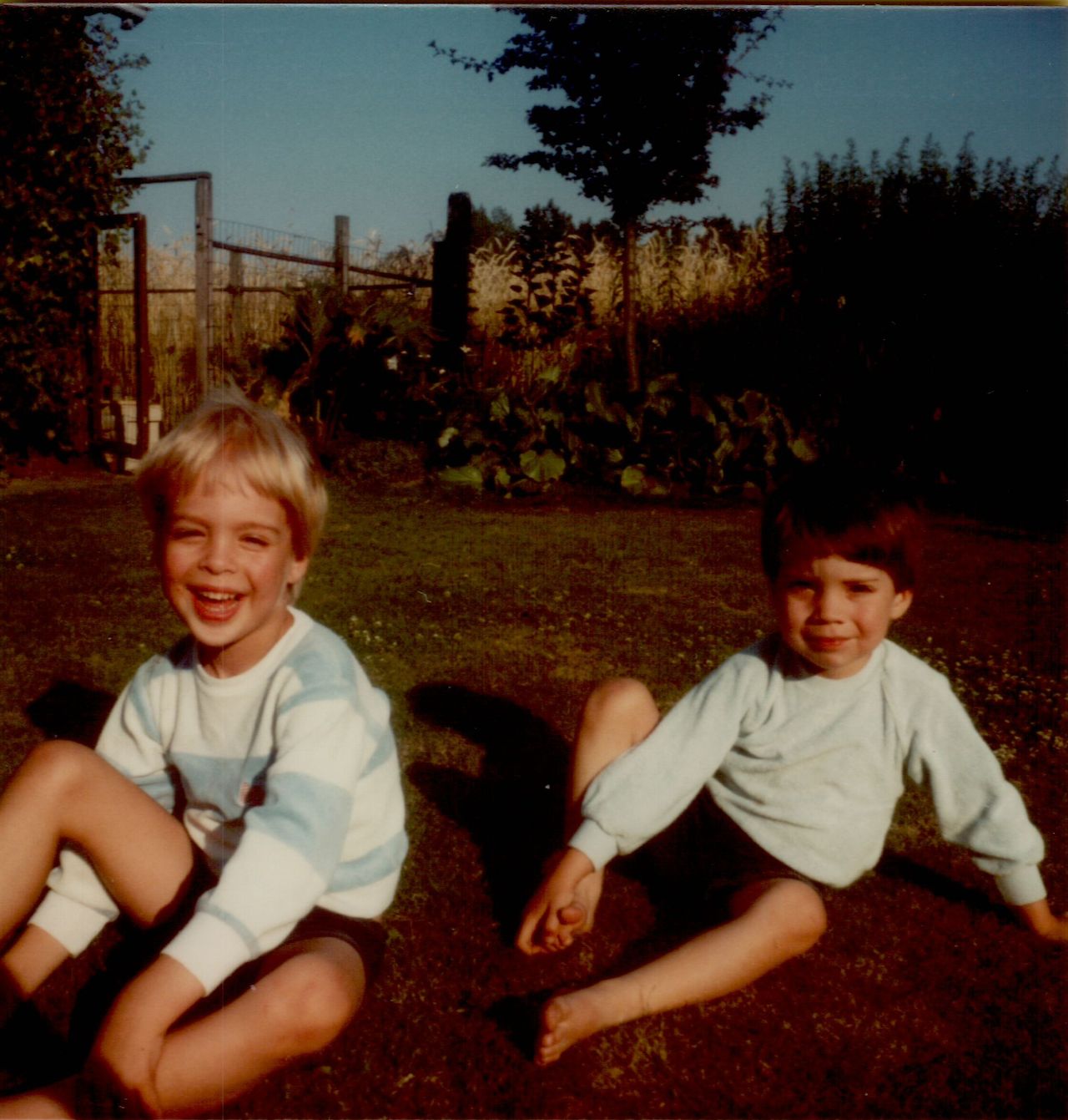 Two young children sit on the grass, smiling and looking at the camera on a sunny day.