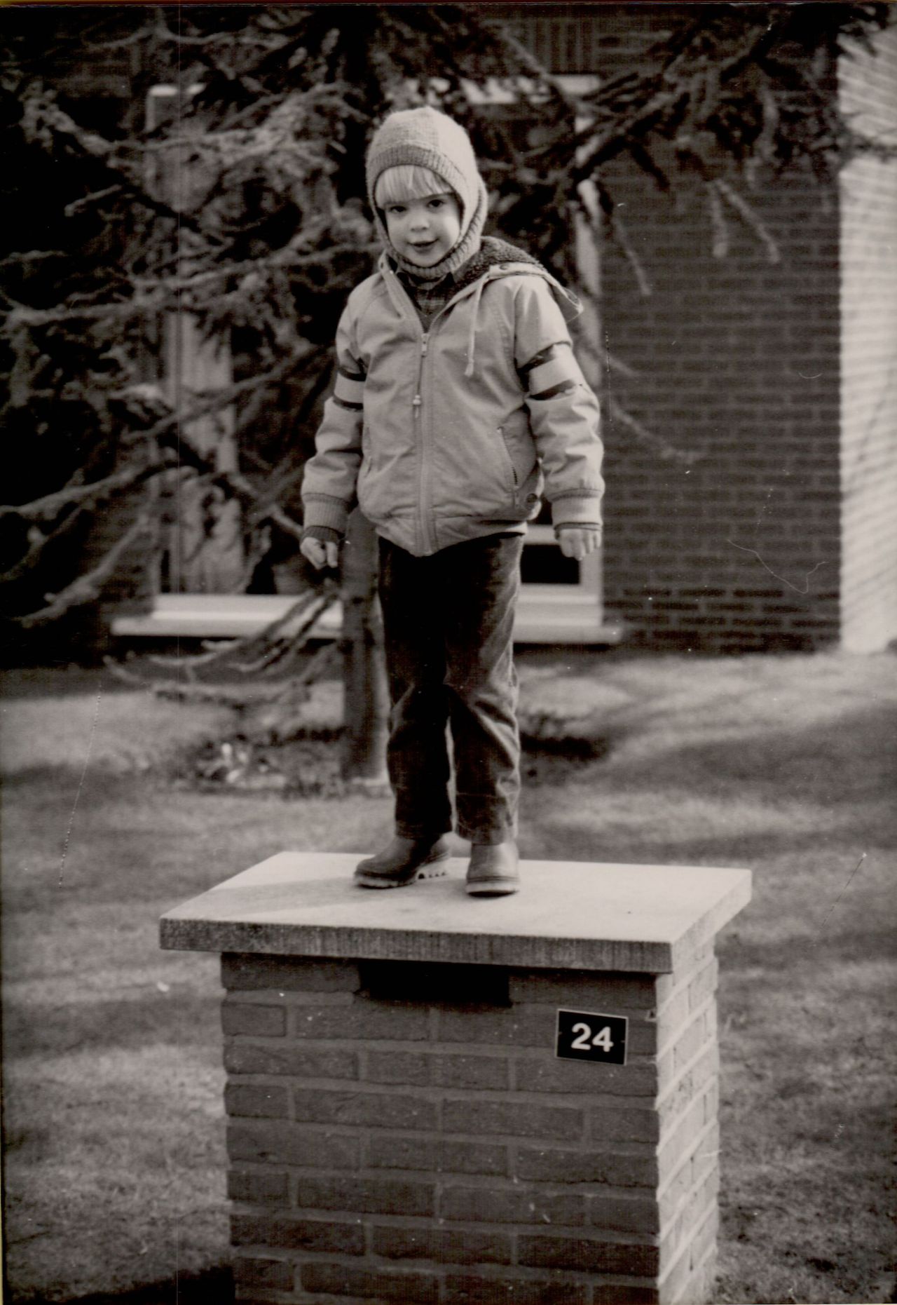 A child wearing a winter jacket and hat stands on top of a brick mailbox outside a house.