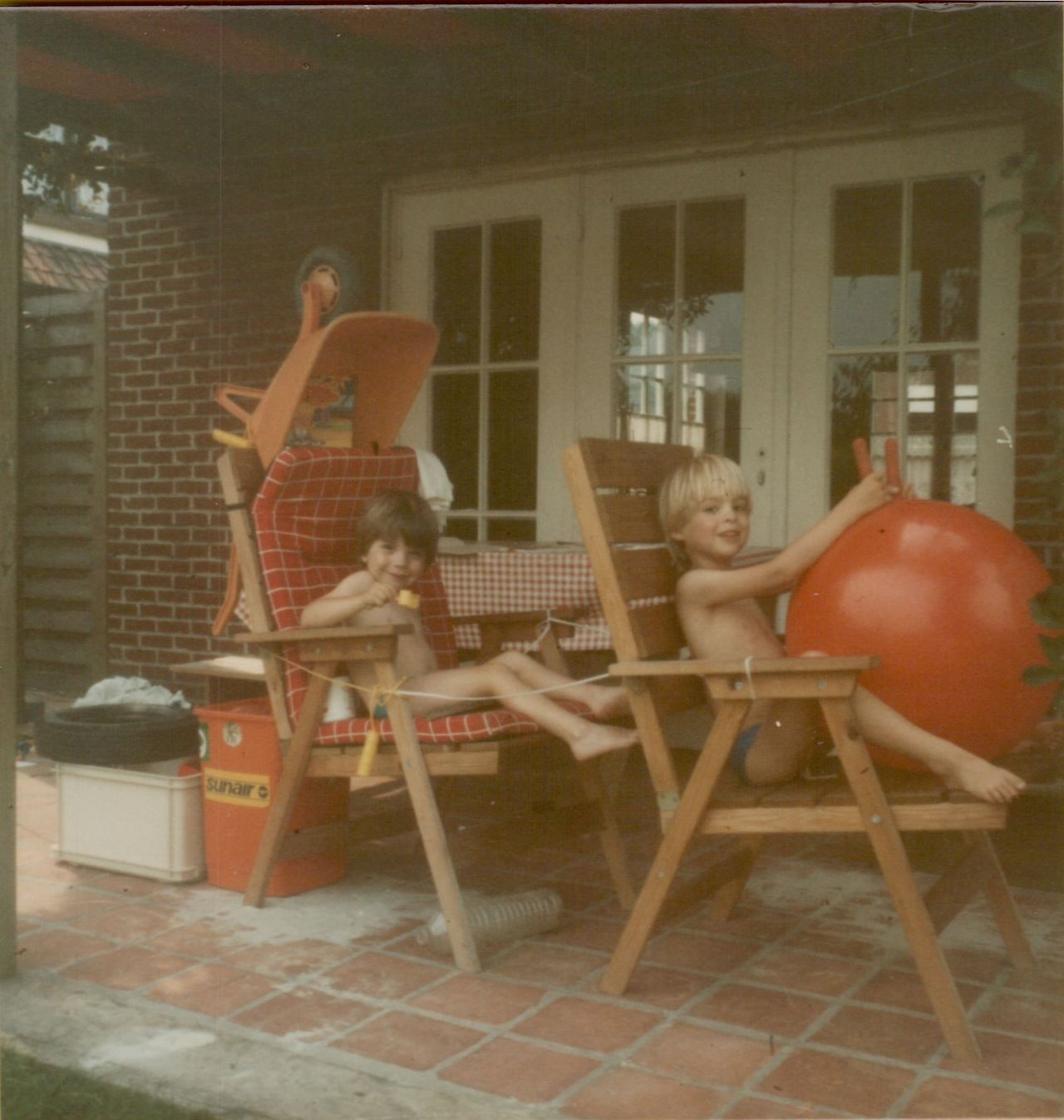 Two children sit on wooden chairs, pretending to drive using a red bouncy ball as a steering wheel.