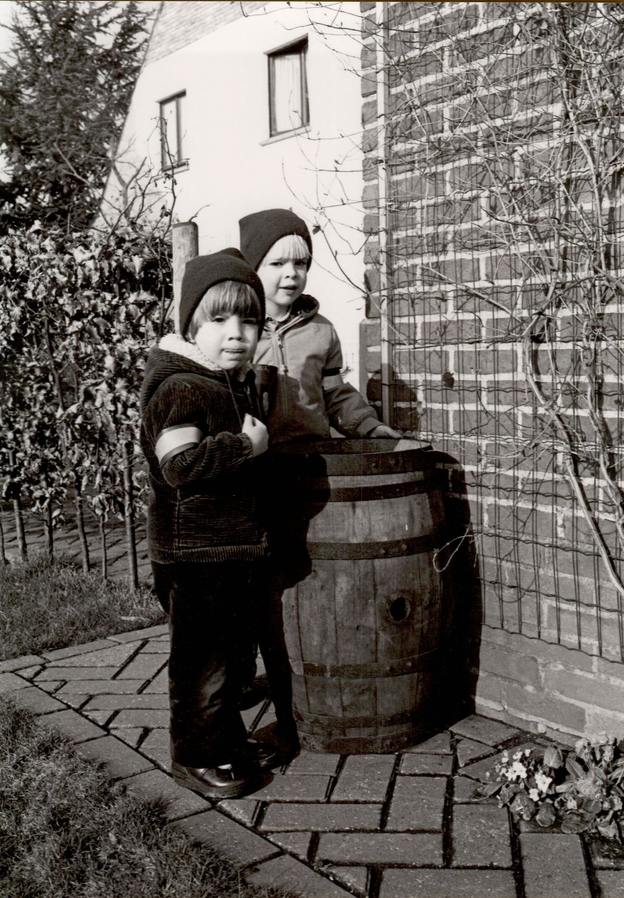 Two children wearing winter clothes stand next to a wooden barrel, pretending to make soup outside.