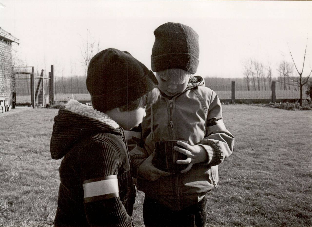 Two children wearing winter jackets and hats stand outside, looking into a cup as if making soup.