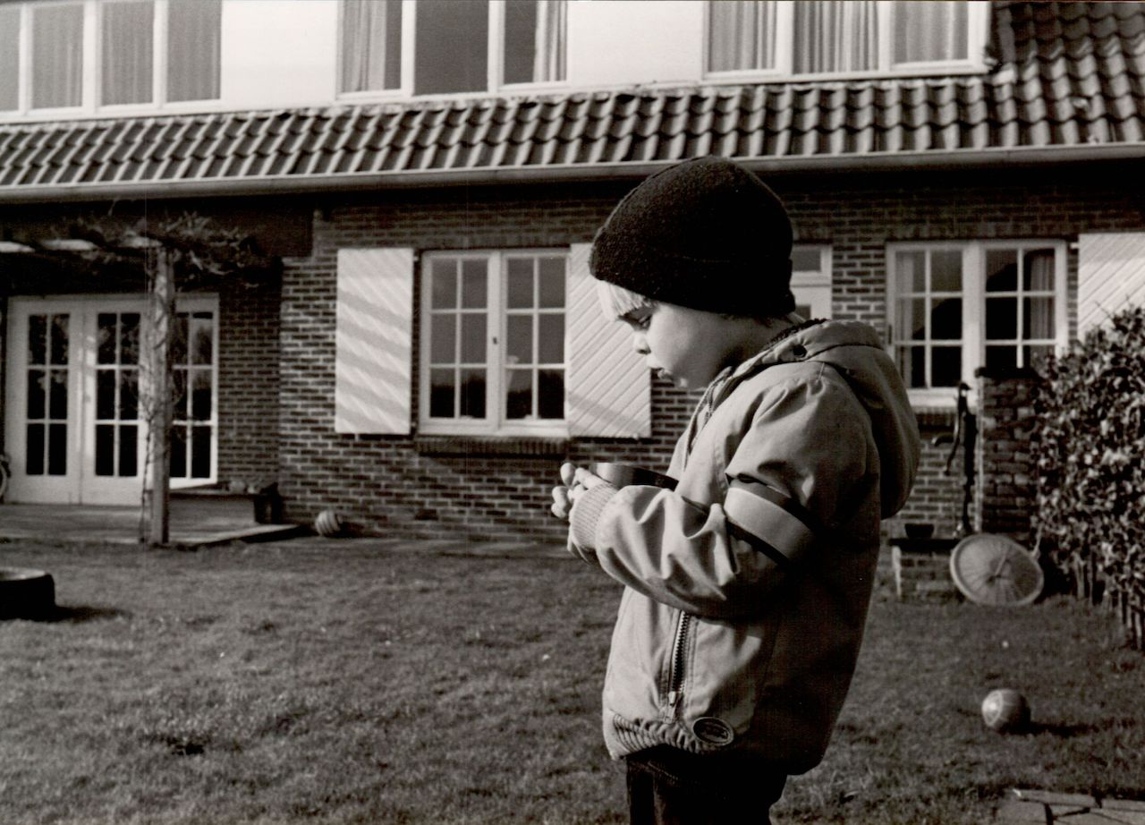 A child in a jacket and beanie looks down at a small cup while standing outside a house.