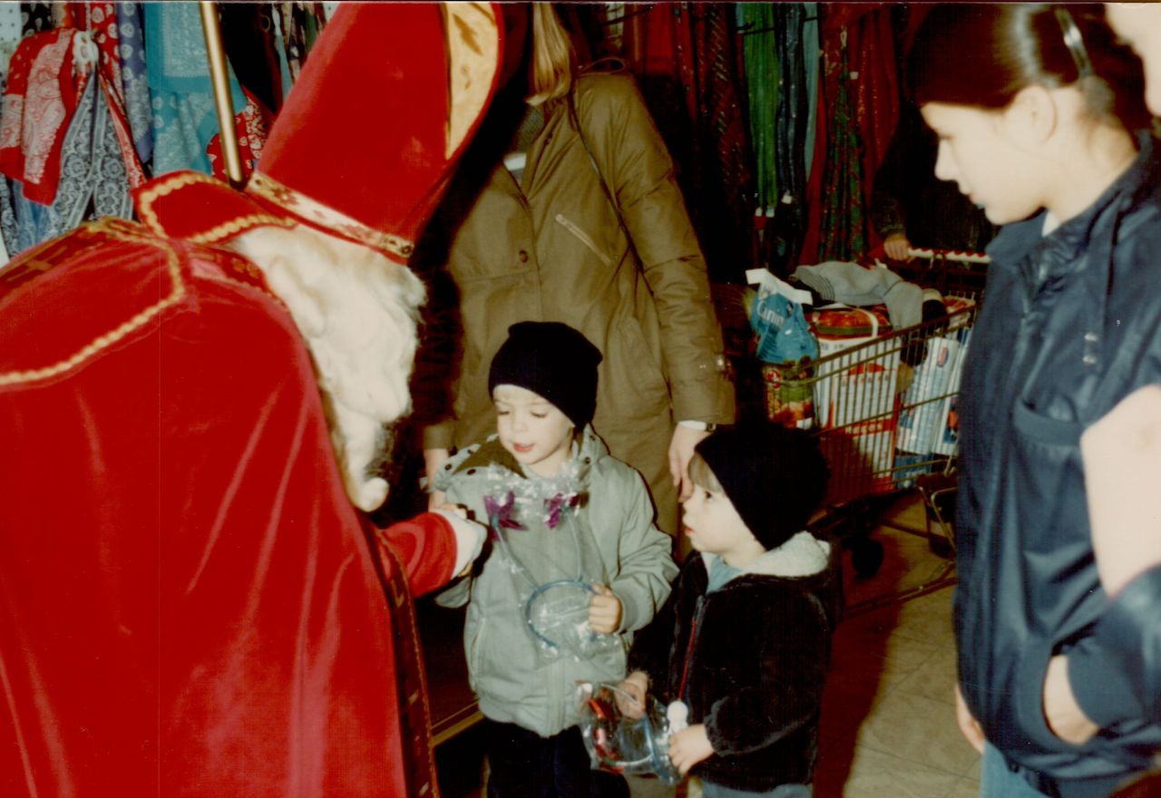 Sinterklaas in a red robe hands a gift to a child while another child watches in a busy store.
