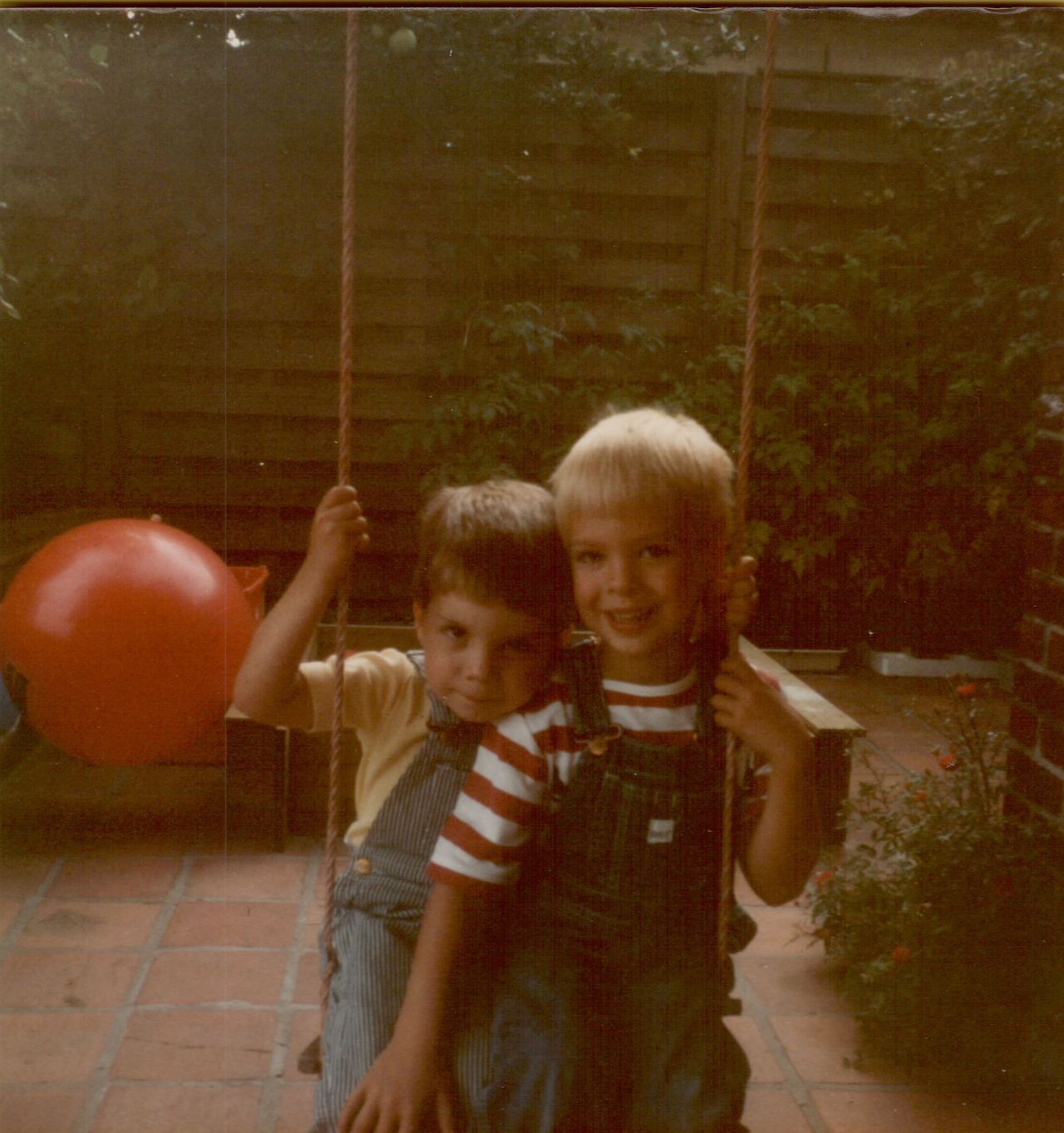 Two young children sit together on a swing, holding the ropes and smiling.