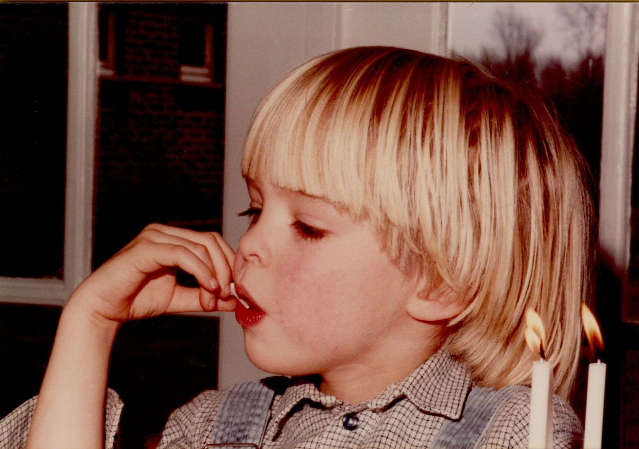 A young child with blonde hair licks frosting off their finger near two lit birthday candles.