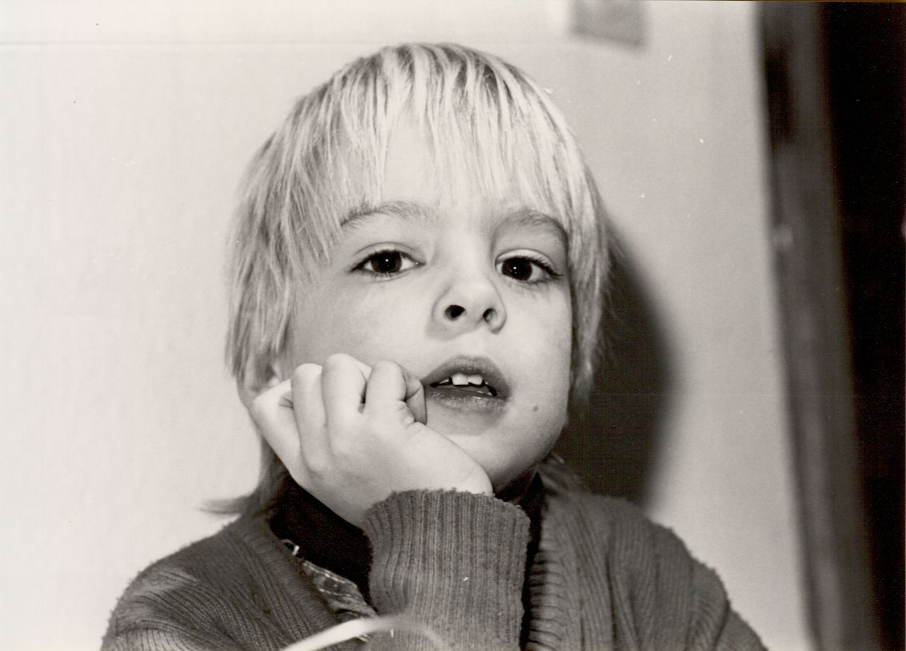 A young child with light hair rests their chin on their hand, looking directly at the camera.