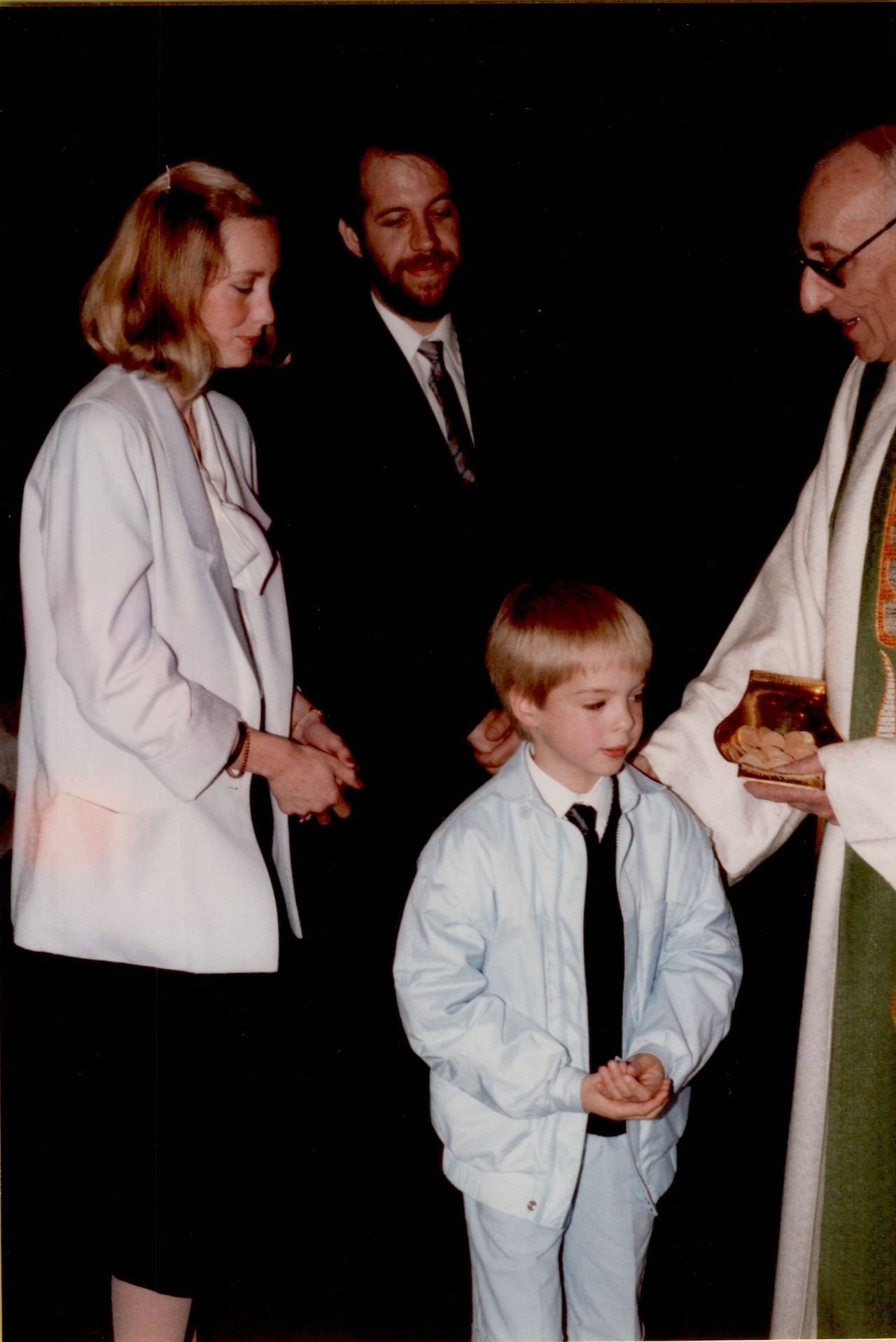 A young boy in a light suit receives communion from a priest, with two adults standing nearby.