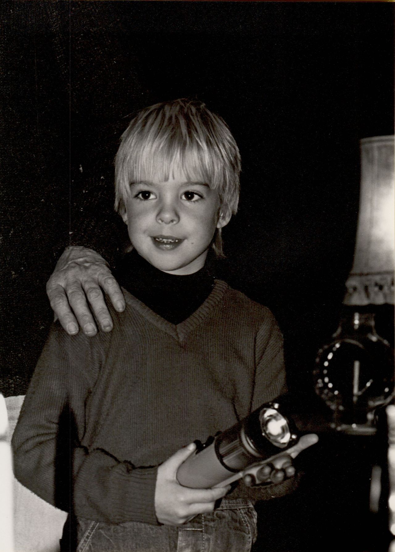 A young child holds a large flashlight, smiling, with an adult's hand resting on their shoulder.
