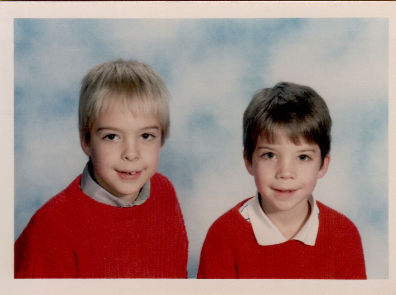 Two young boys wearing red sweaters pose for a school photo against a blue and white background.