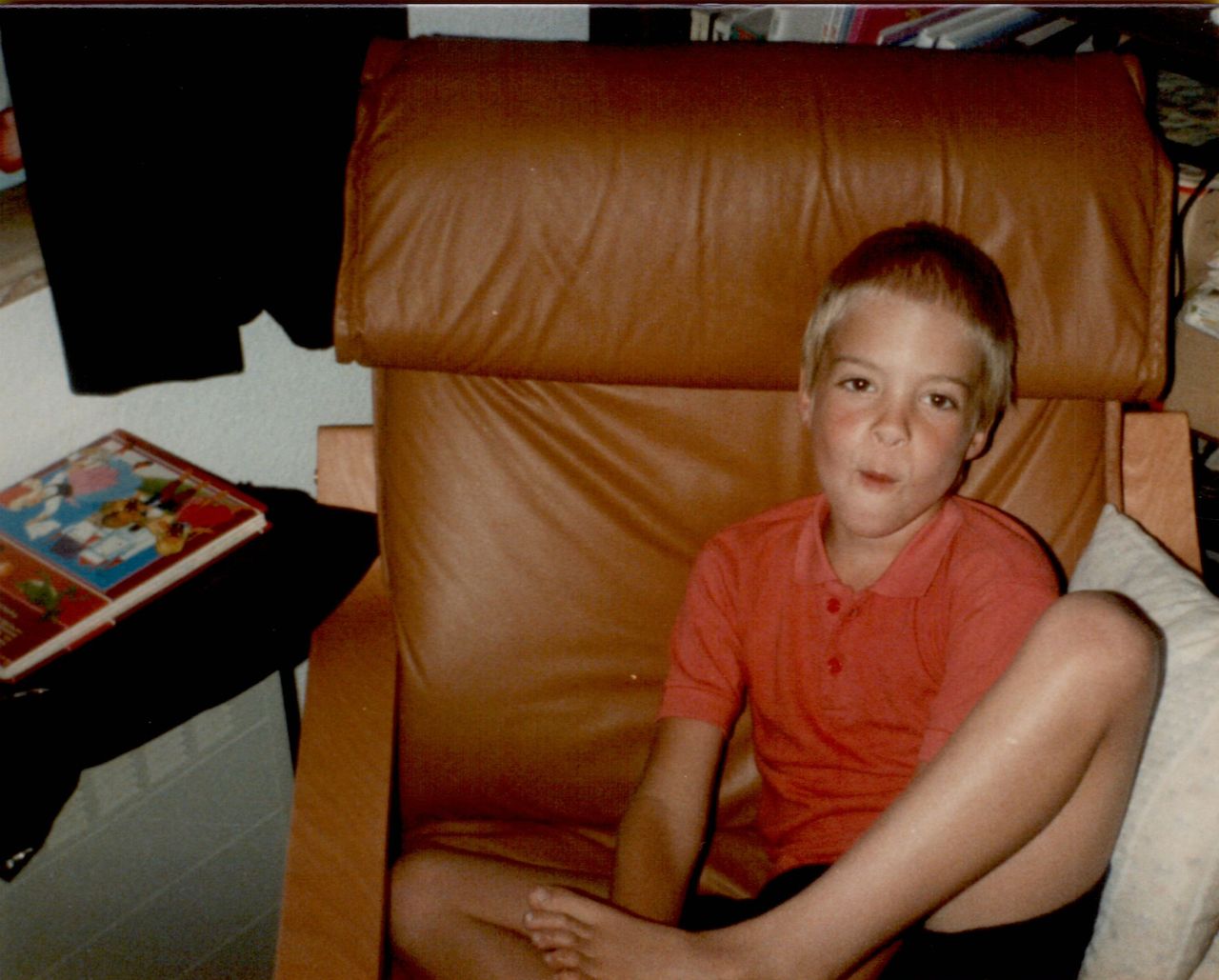 A young boy in a red shirt sits in a brown IKEA chair, making a playful facial expression.