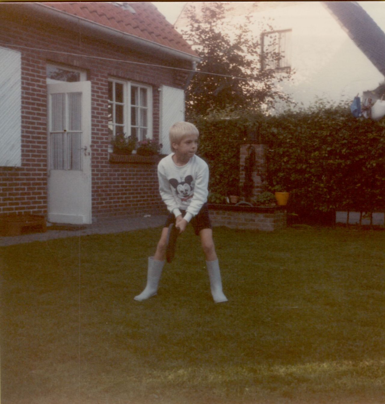 A young child in a sweatshirt and boots holds a baseball bat, preparing to swing in a grassy backyard.