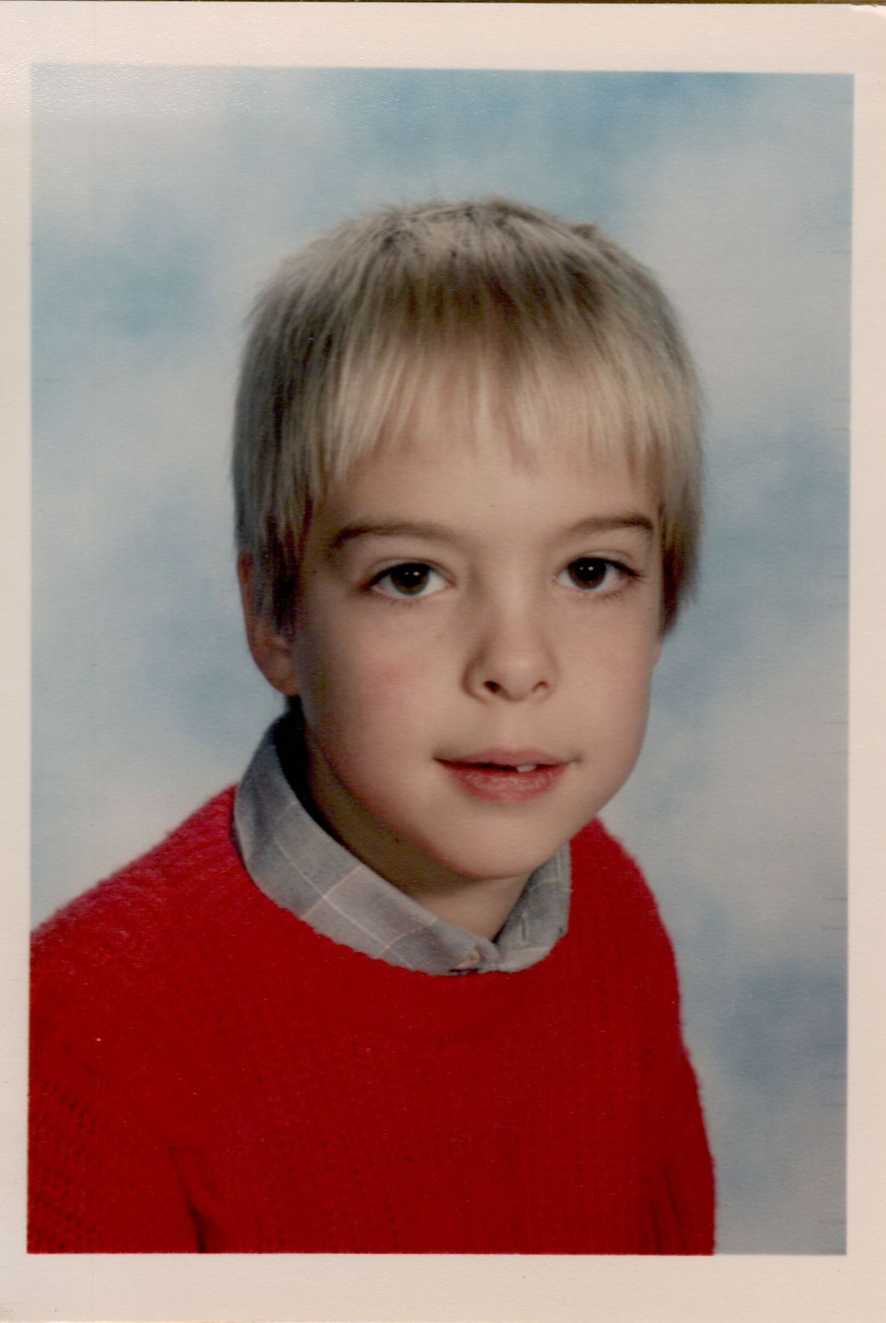A young child with short blond hair wears a red sweater and poses for a school photo against a blue background.
