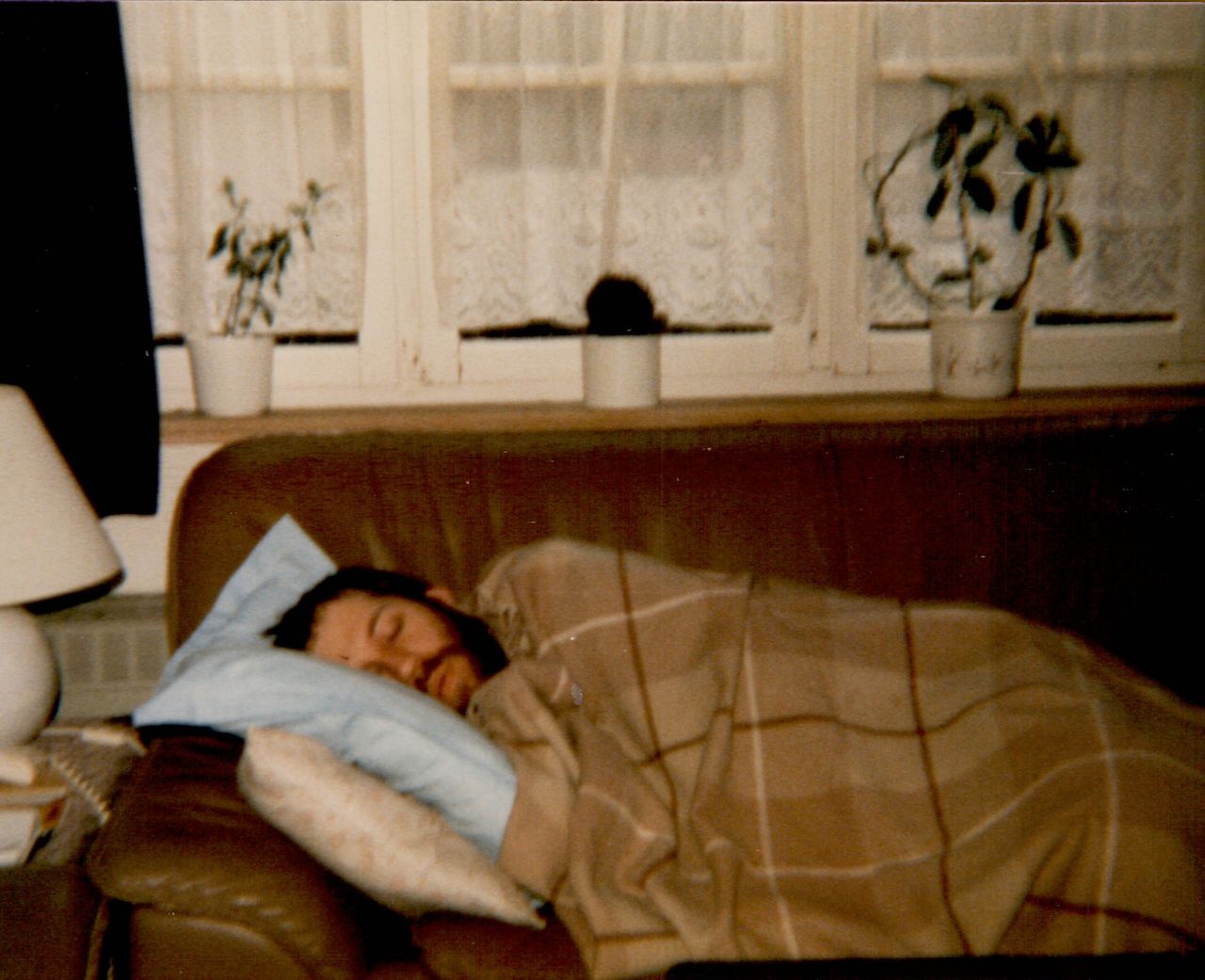 A man with a beard is sleeping on a couch, covered with a blanket and resting on multiple pillows.