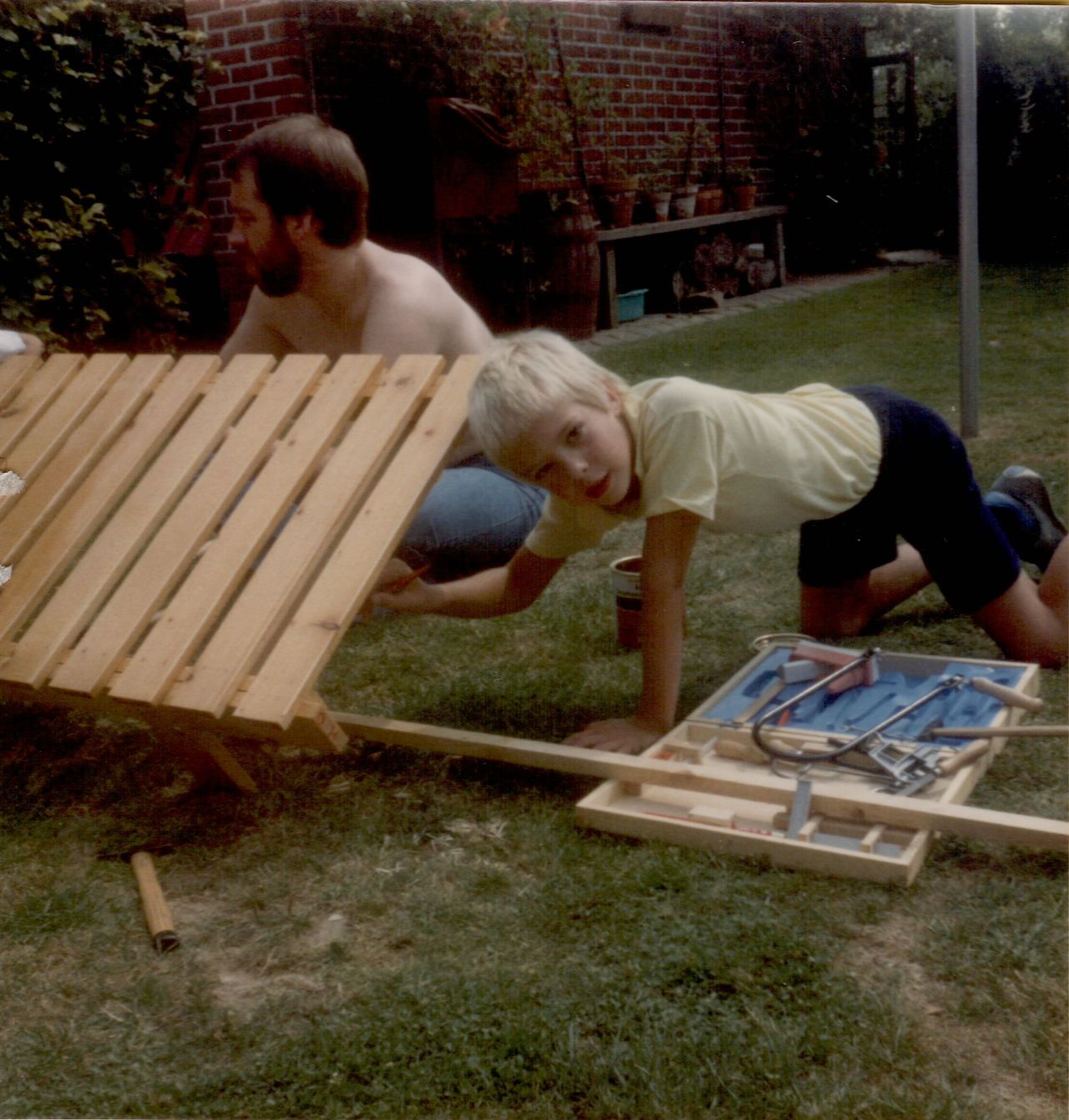 A man and a child assemble a wooden sunbed in a grassy backyard, with tools scattered nearby.