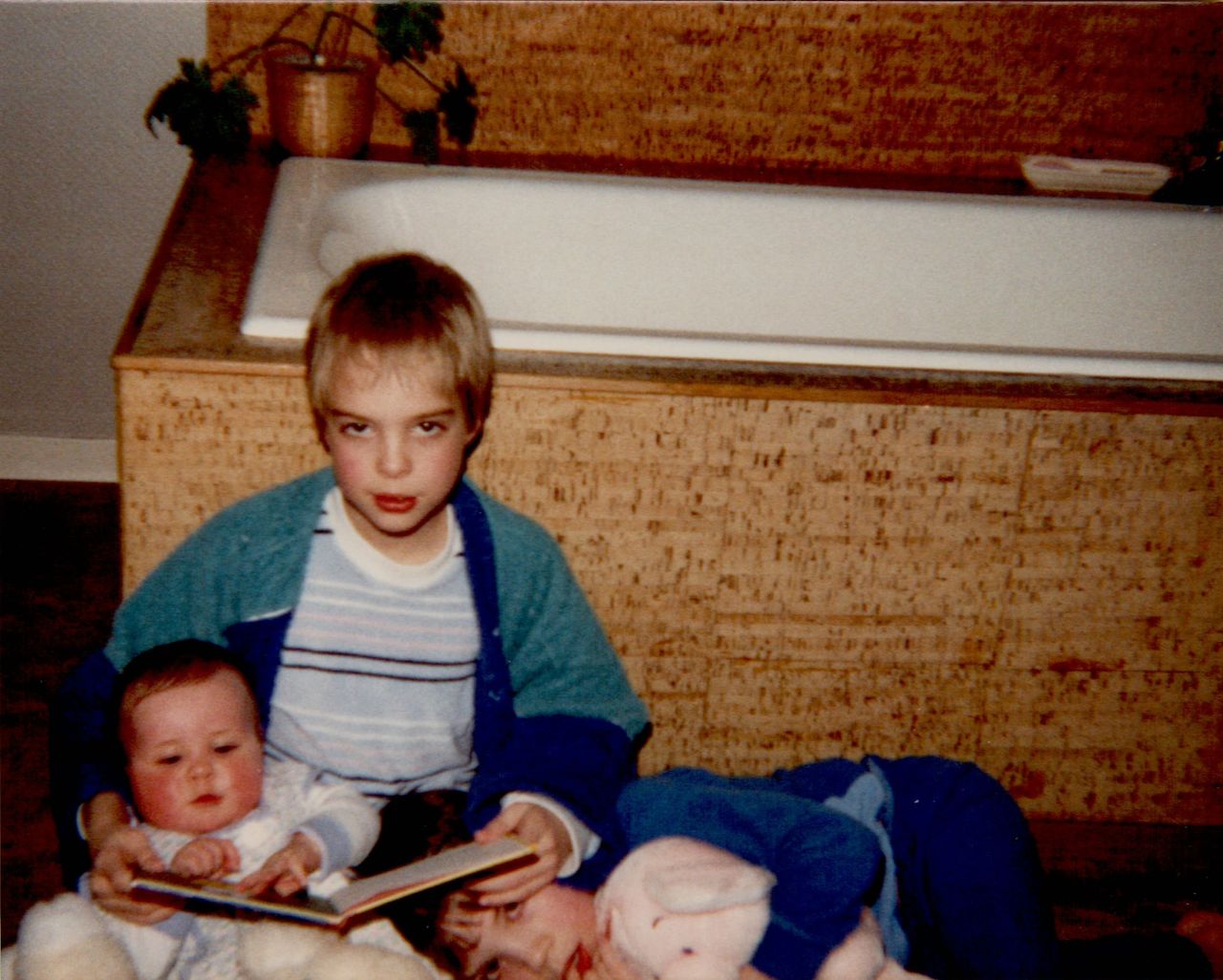 A child sits holding a book, reading to a baby and another sibling lying down with a stuffed animal.