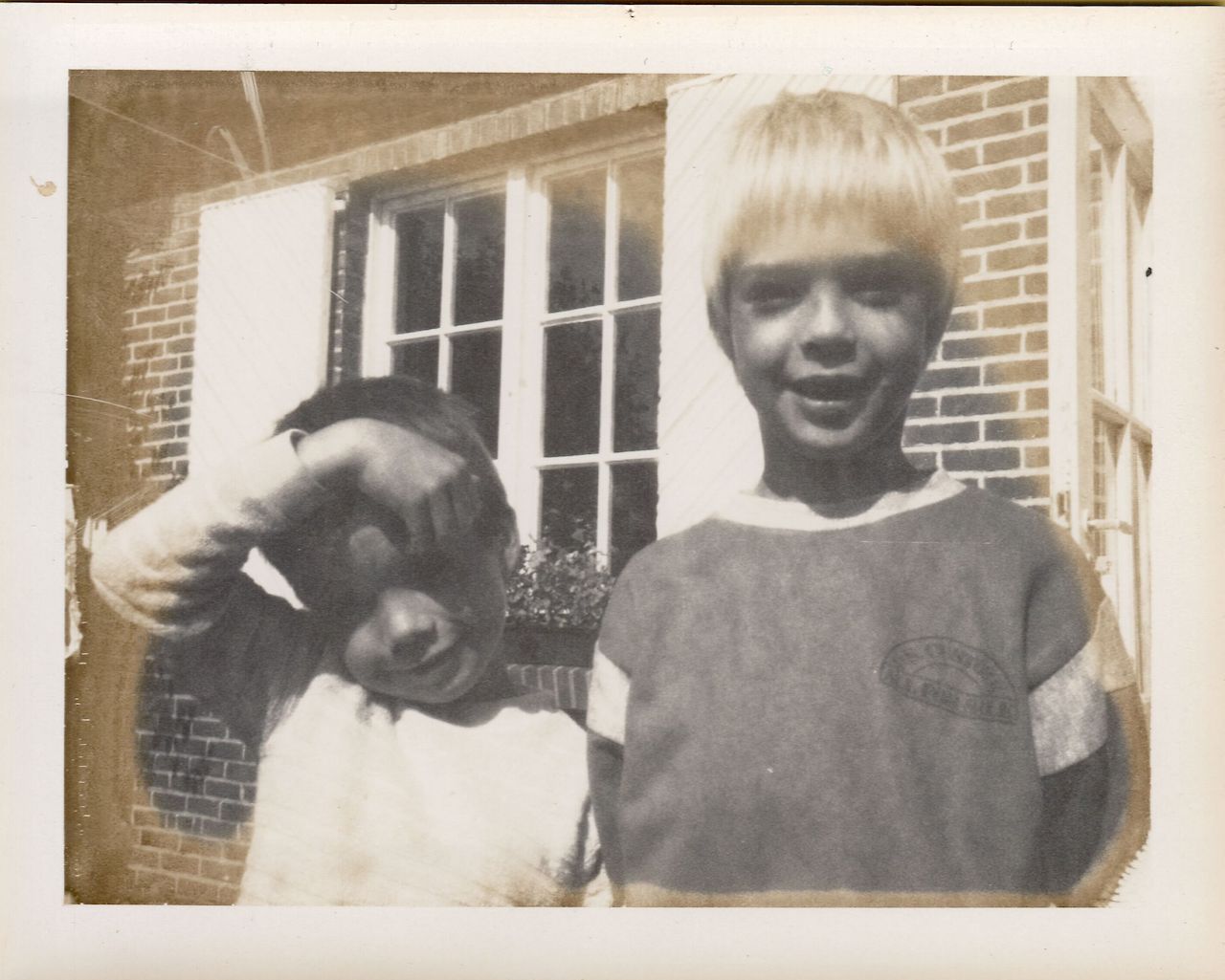 Two young boys stand outside a brick house, one smiling and the other shielding his eyes from the sun.