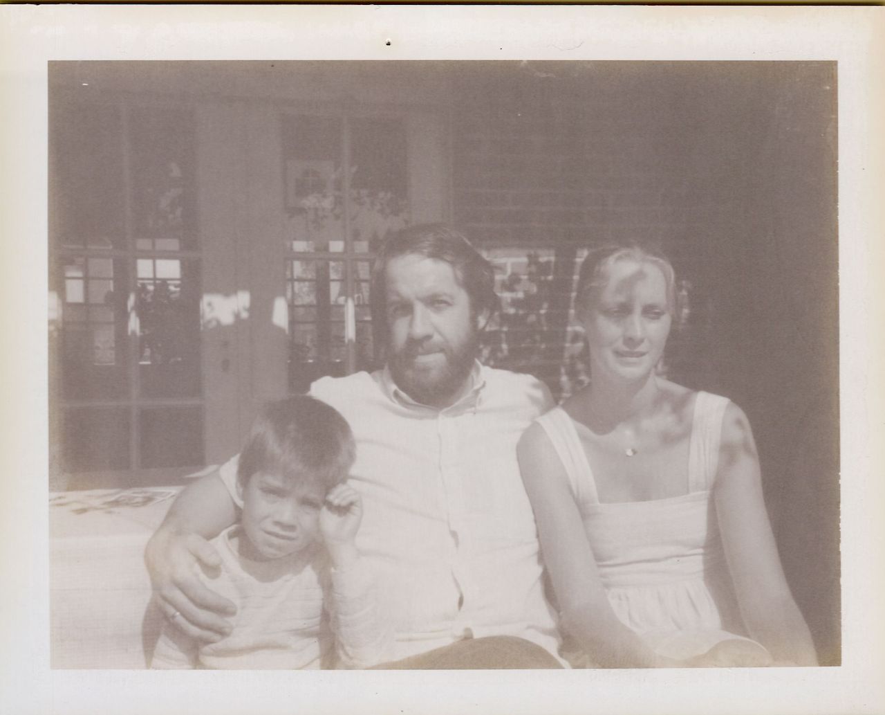 A man sits with his arm around a young child, while a woman sits beside them, all facing the camera.