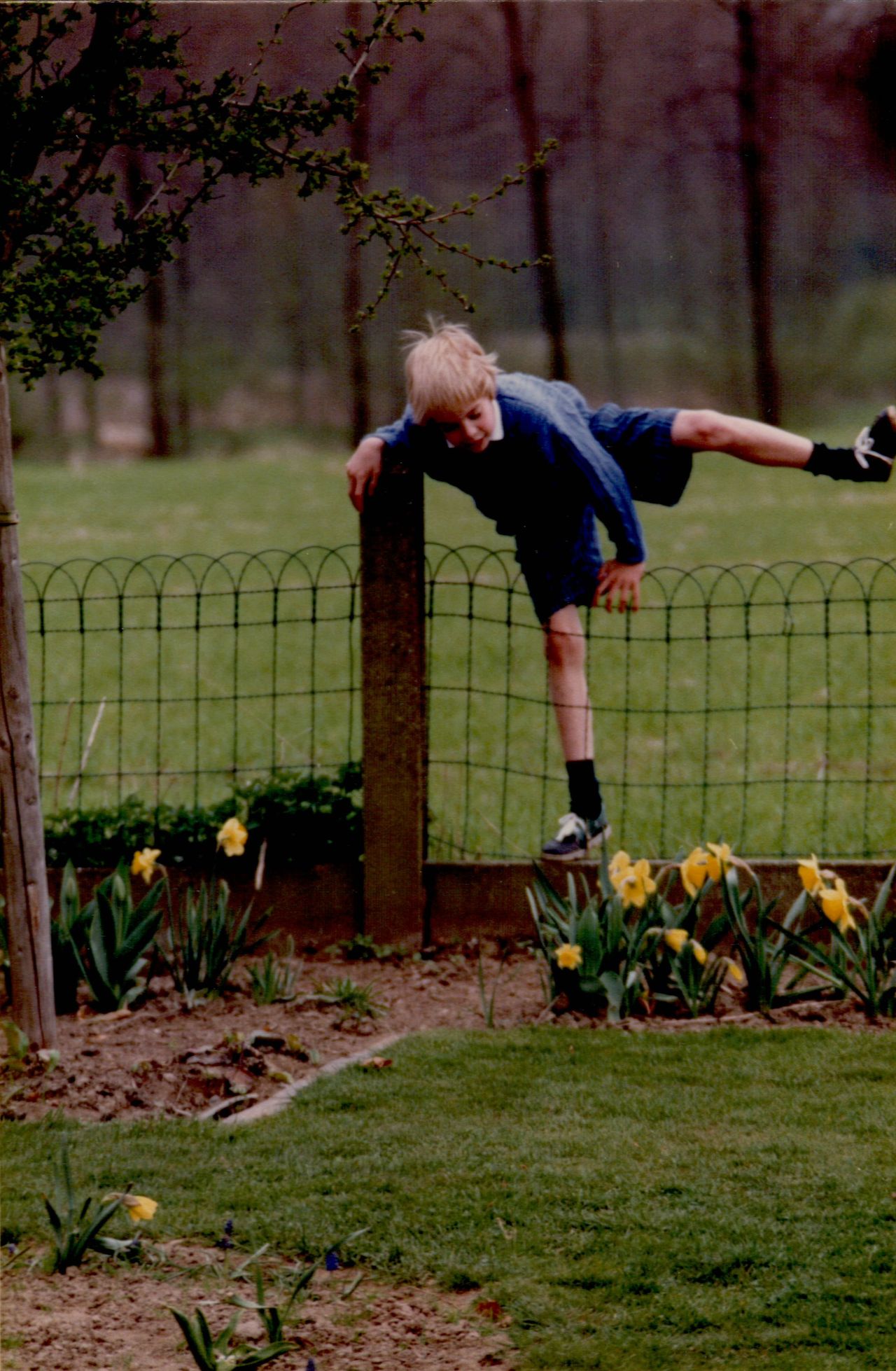 A young boy in a blue outfit climbs over a wire fence, balancing with one leg over the top.