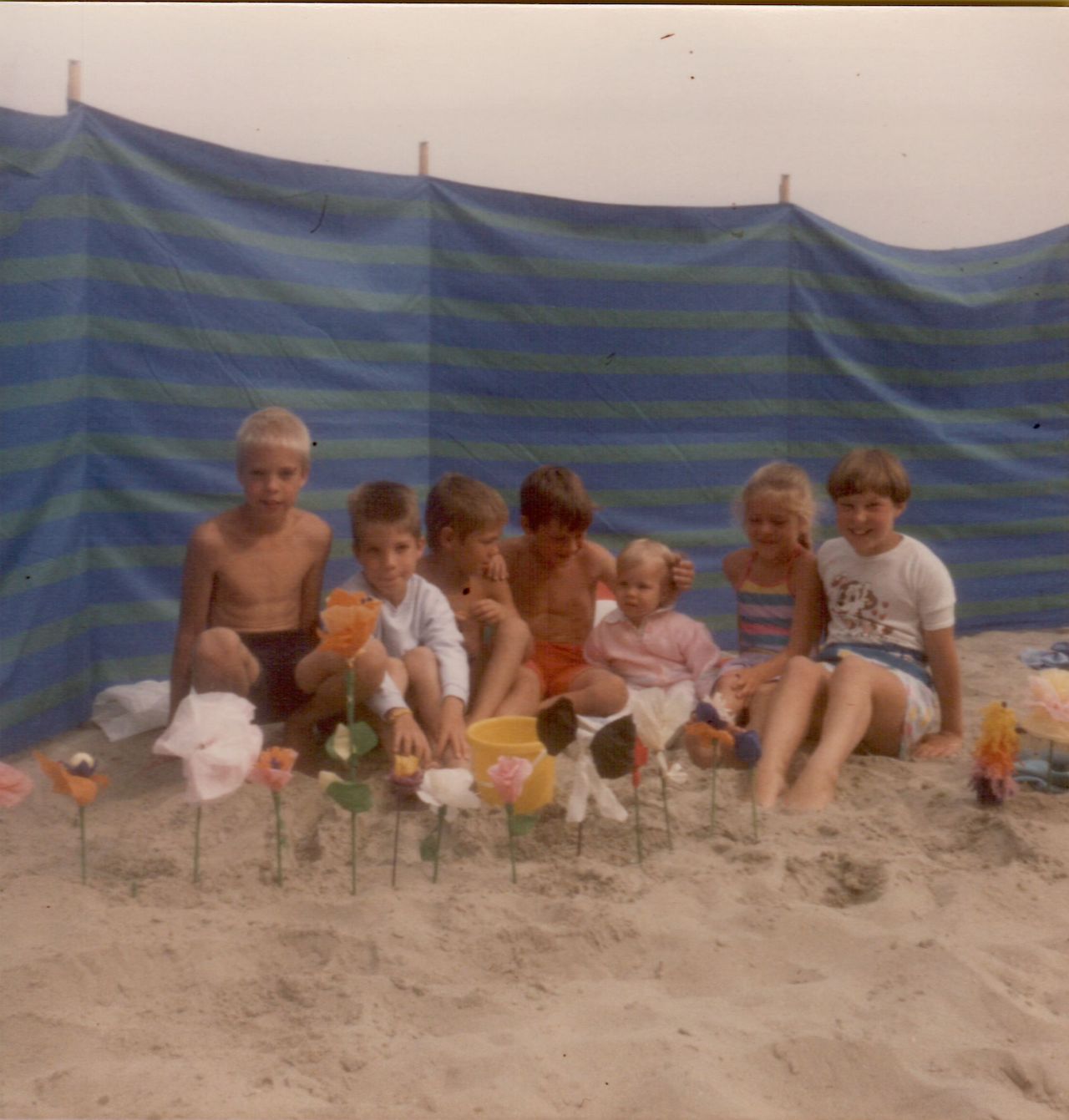 A group of children sits on the sand, smiling and posing behind handmade paper flowers at the beach.