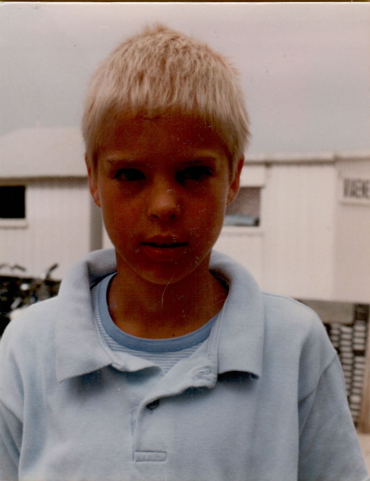 A young child with short blond hair wearing a light blue polo shirt looks directly at the camera.
