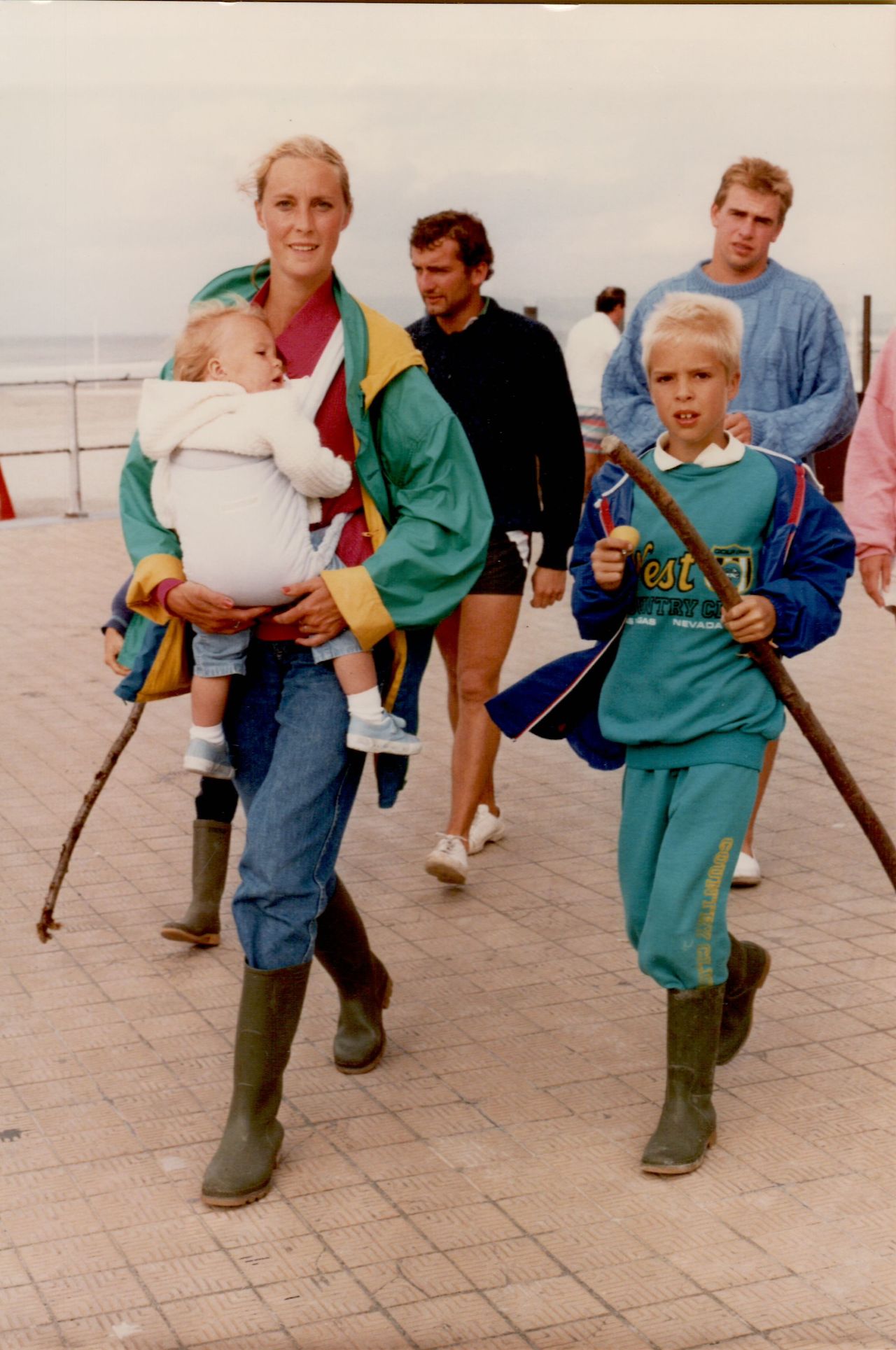 A woman carrying a baby walks alongside a young boy holding a stick on a seaside promenade.