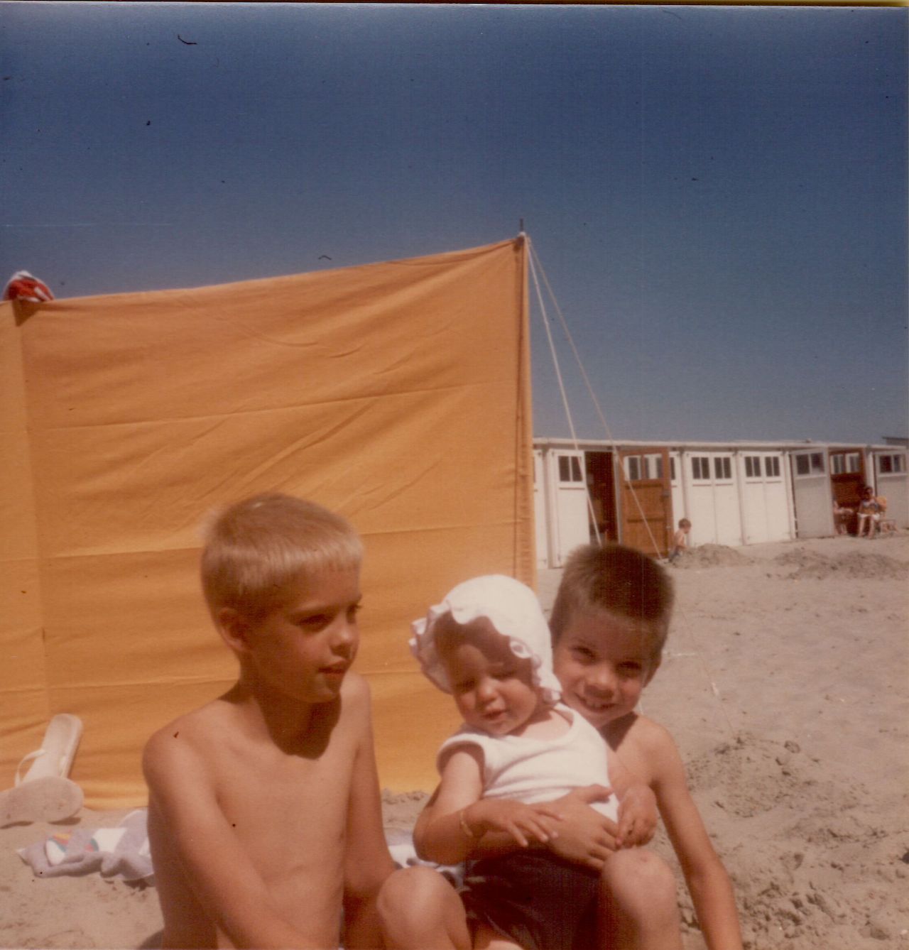 Three children sit on a sandy beach, with one holding a baby wearing a white sun hat.
