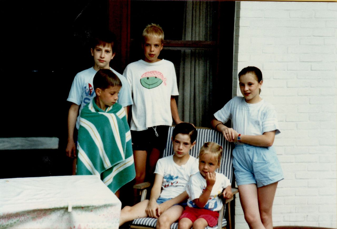 Six children pose together outside a house, with one sitting in a chair holding a younger child on their lap.