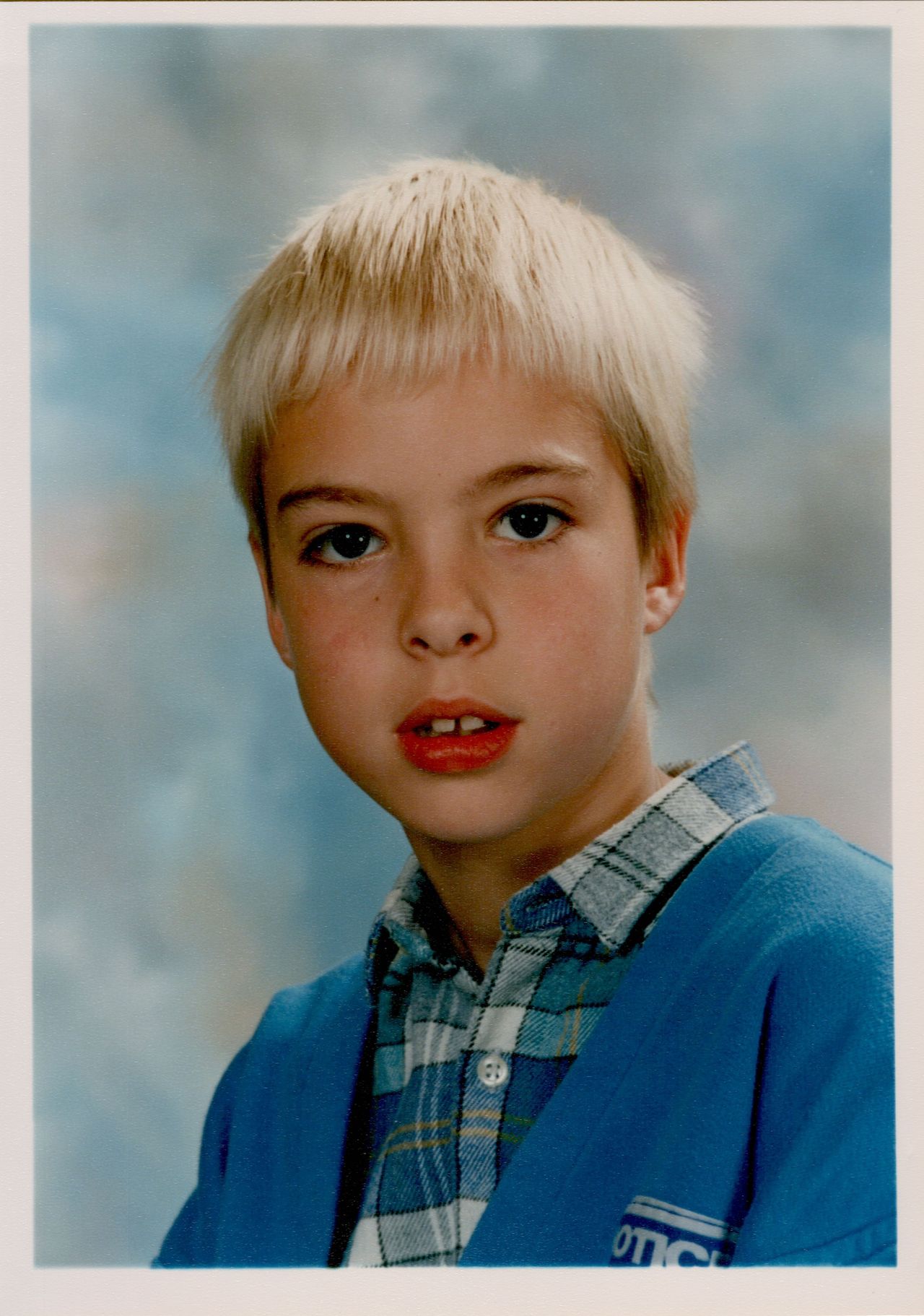A young child with short blond hair poses for a school portrait, wearing a blue sweater over a plaid shirt.