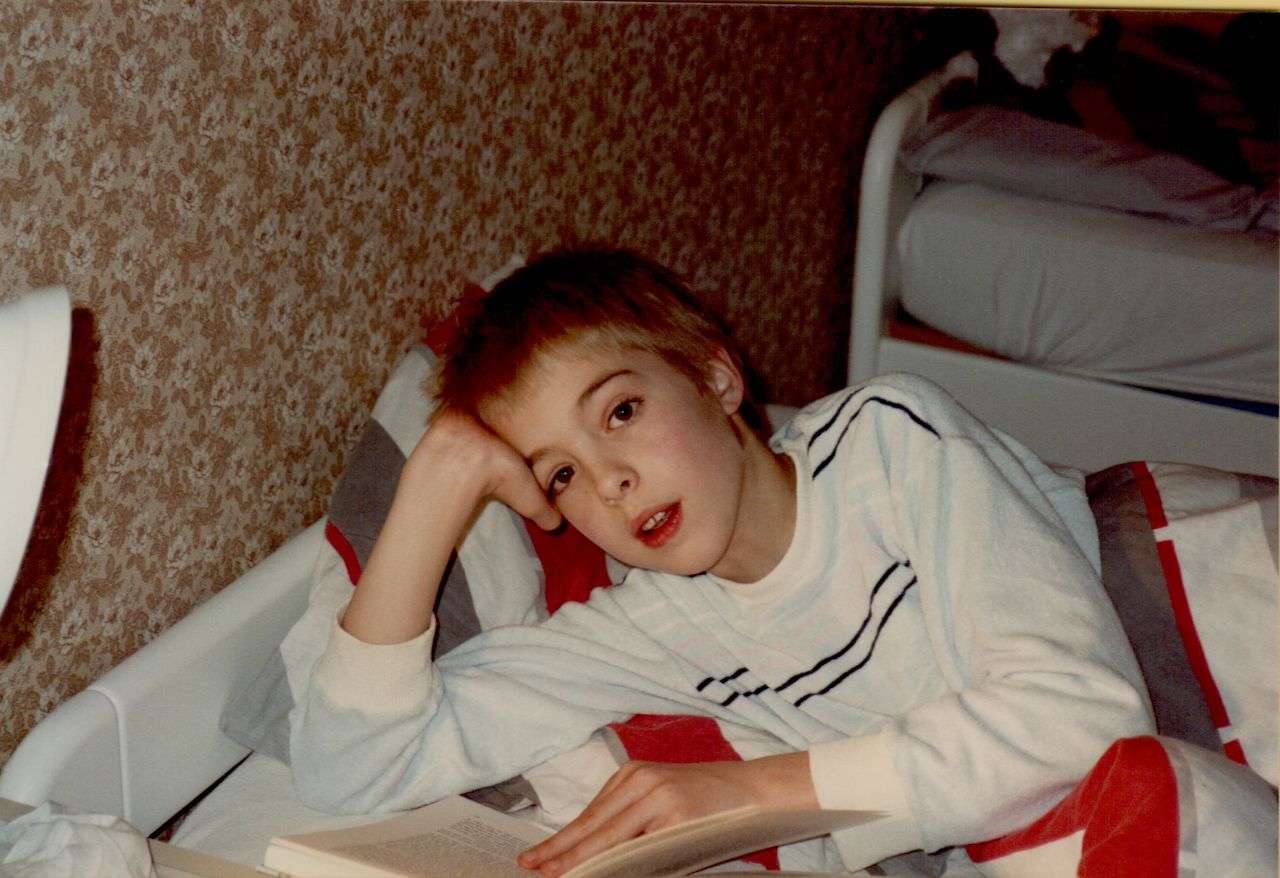 A young person lies in bed, propped on one arm, reading a book with a relaxed expression.