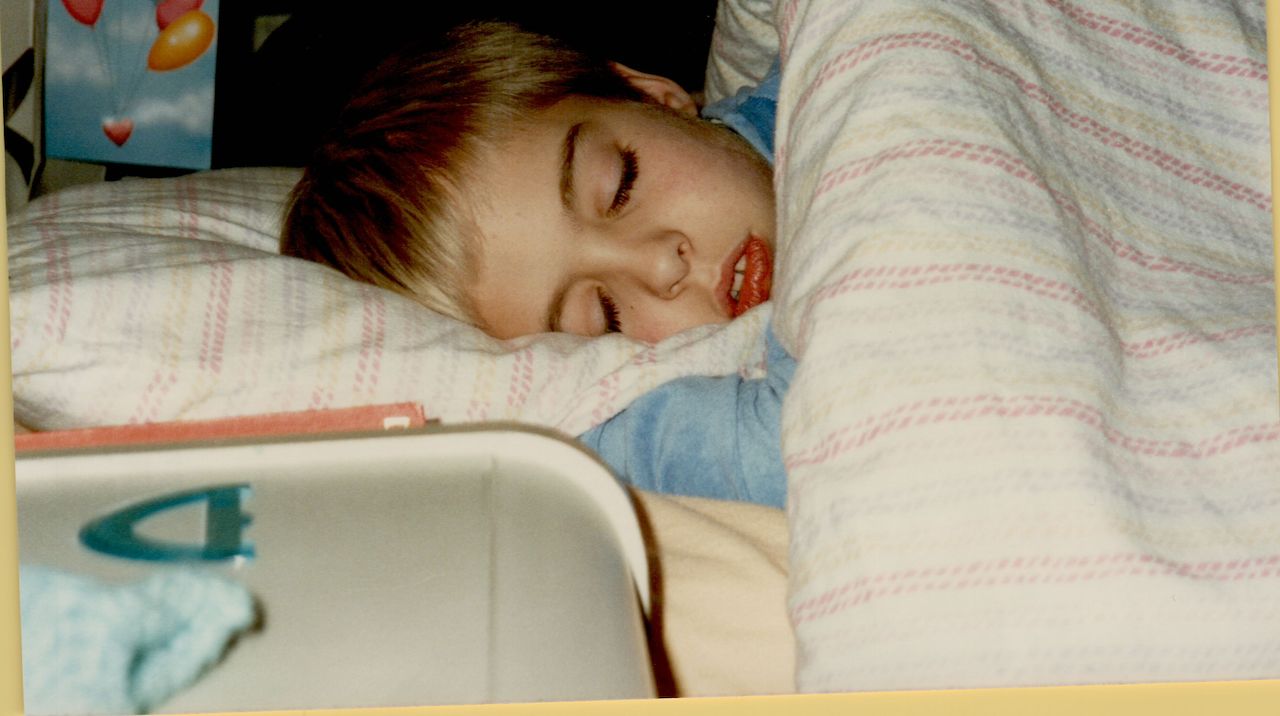 A young child with short blond hair is sleeping under a striped blanket, resting their head on a pillow.