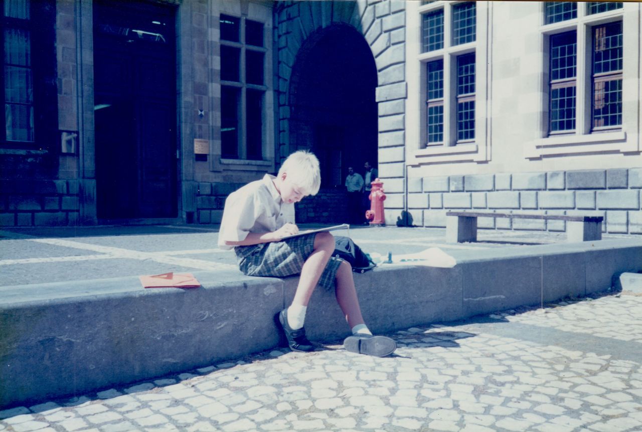 A young person sits on a stone ledge, focused on drawing or writing in a notebook in an outdoor square.