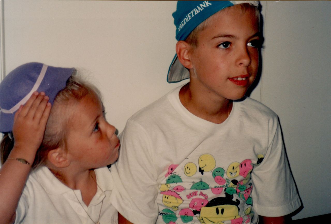 A young girl adjusts her hat while looking at an older boy wearing a backward cap and a colorful shirt.