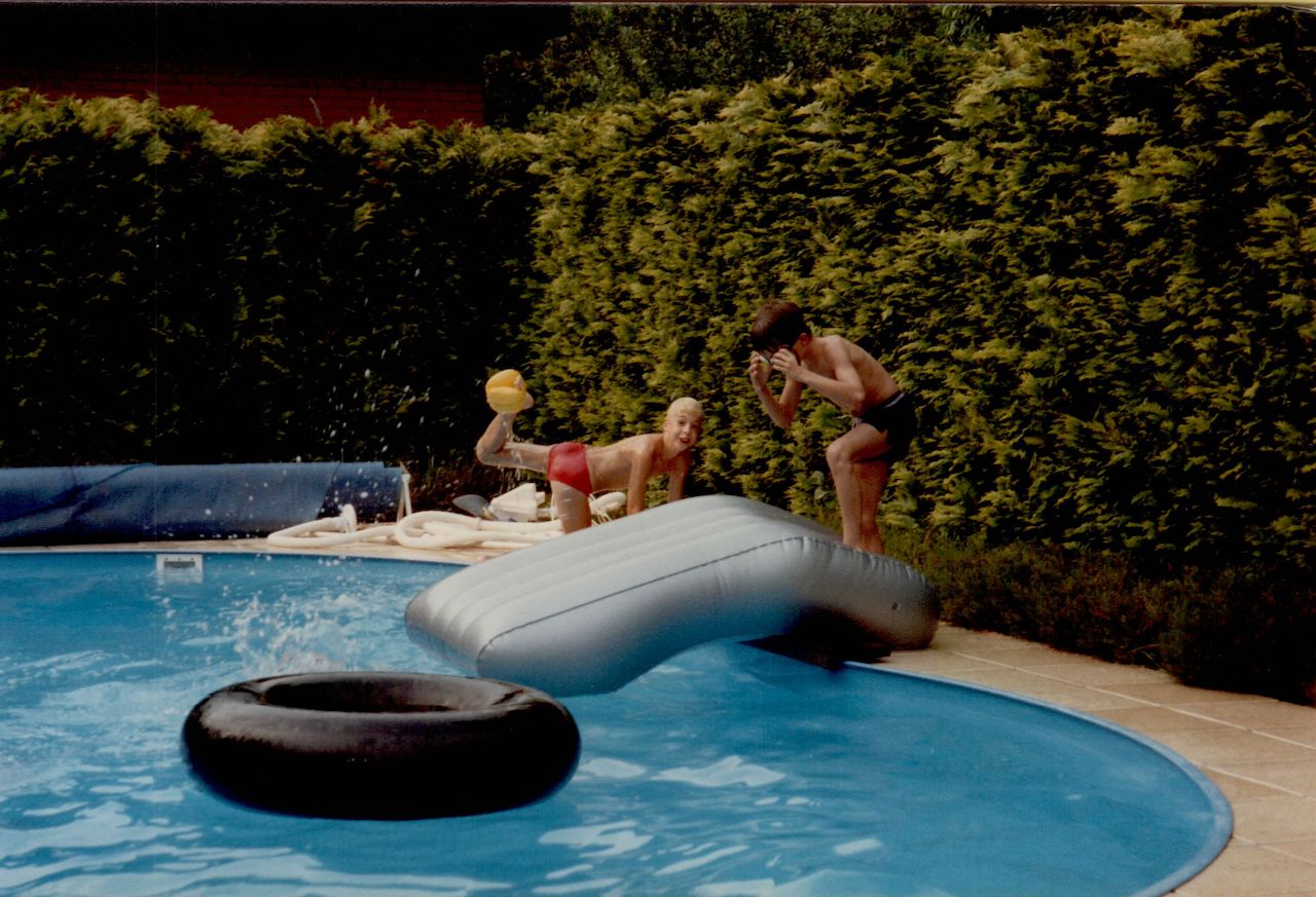 Two children play near a pool, one holding a yellow ball and the other adjusting swimming goggles.