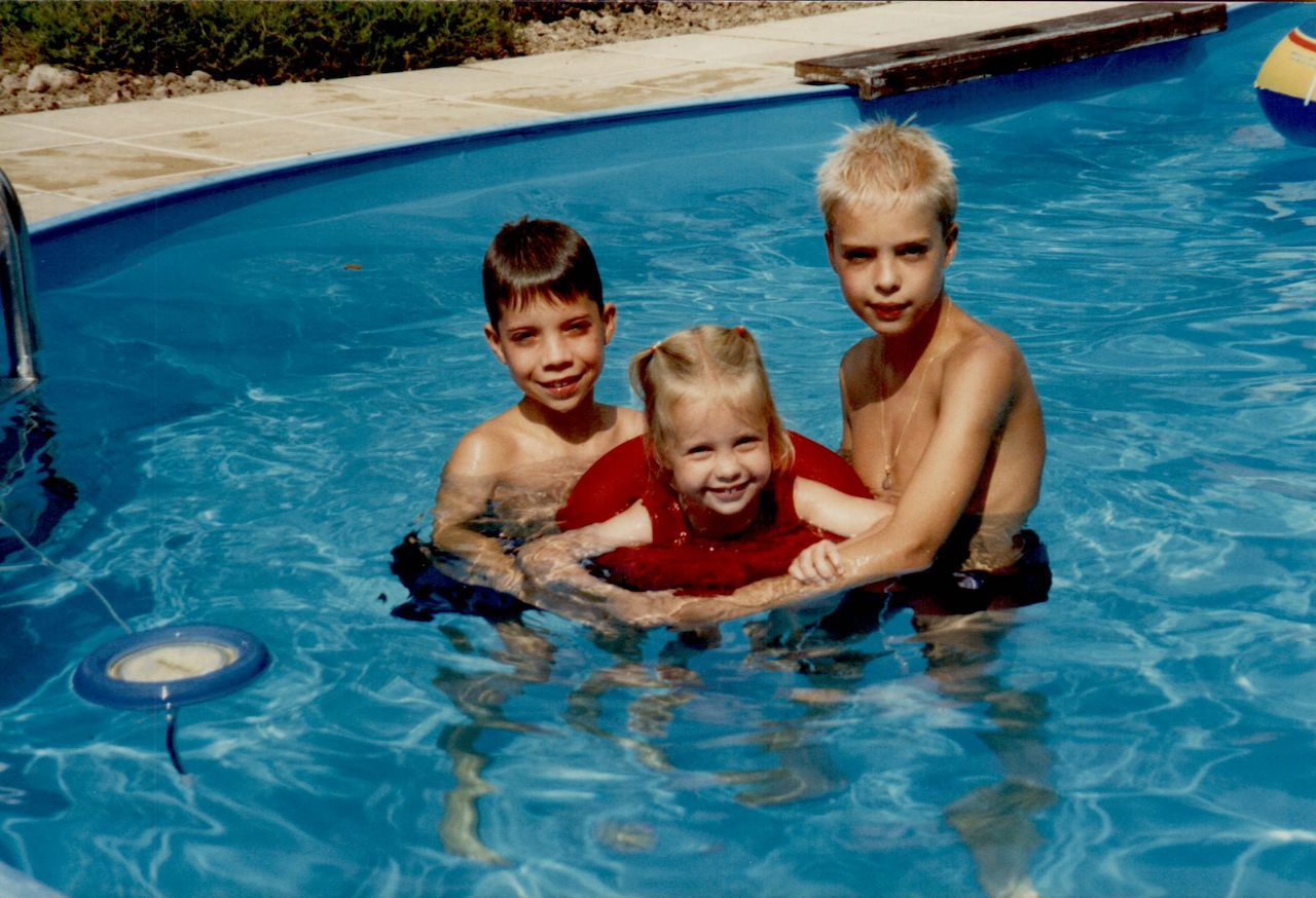 Three children in a swimming pool, with two older boys helping a younger girl float using a red inflatable ring.