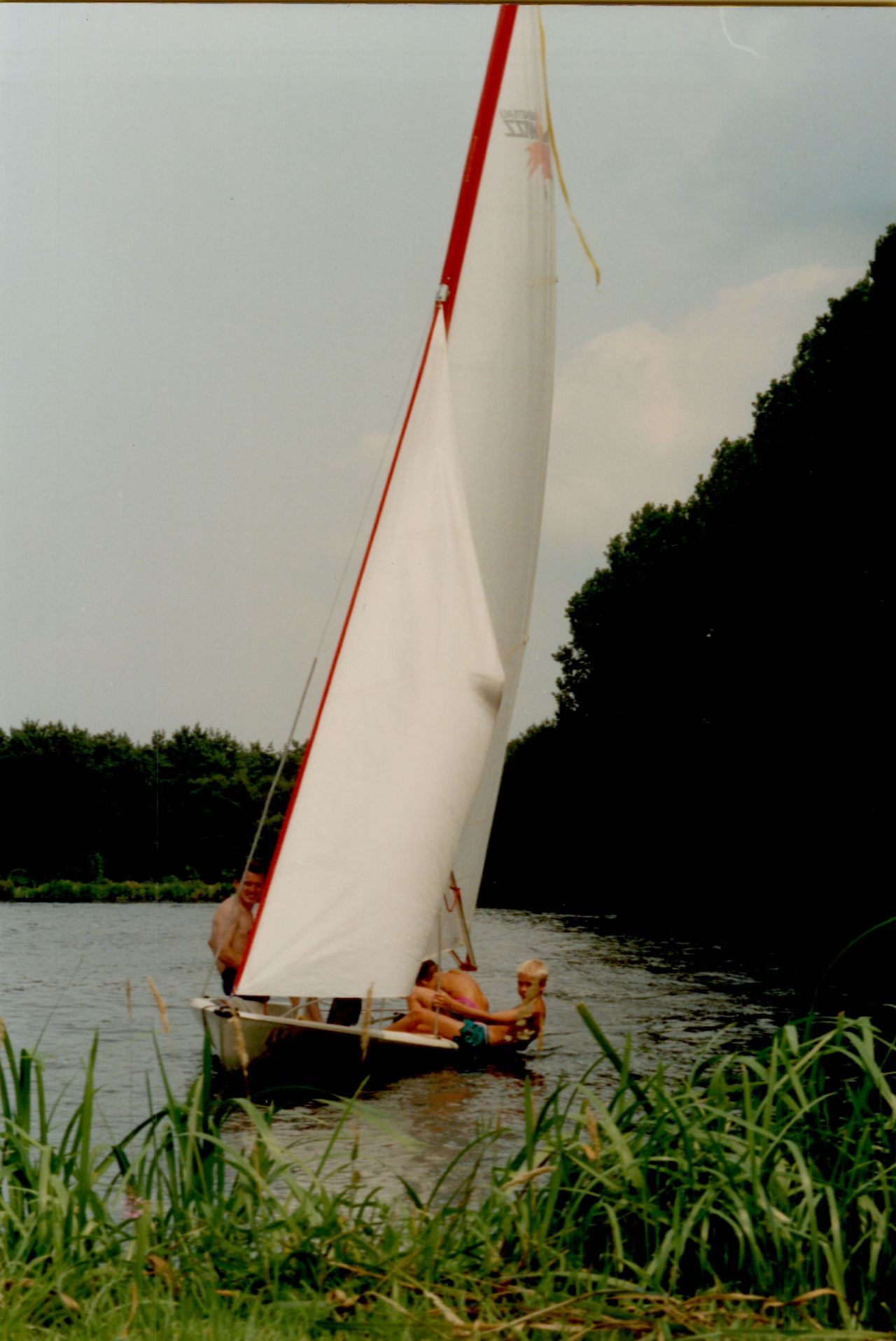 A small sailboat with three people glides on the water, surrounded by greenery.