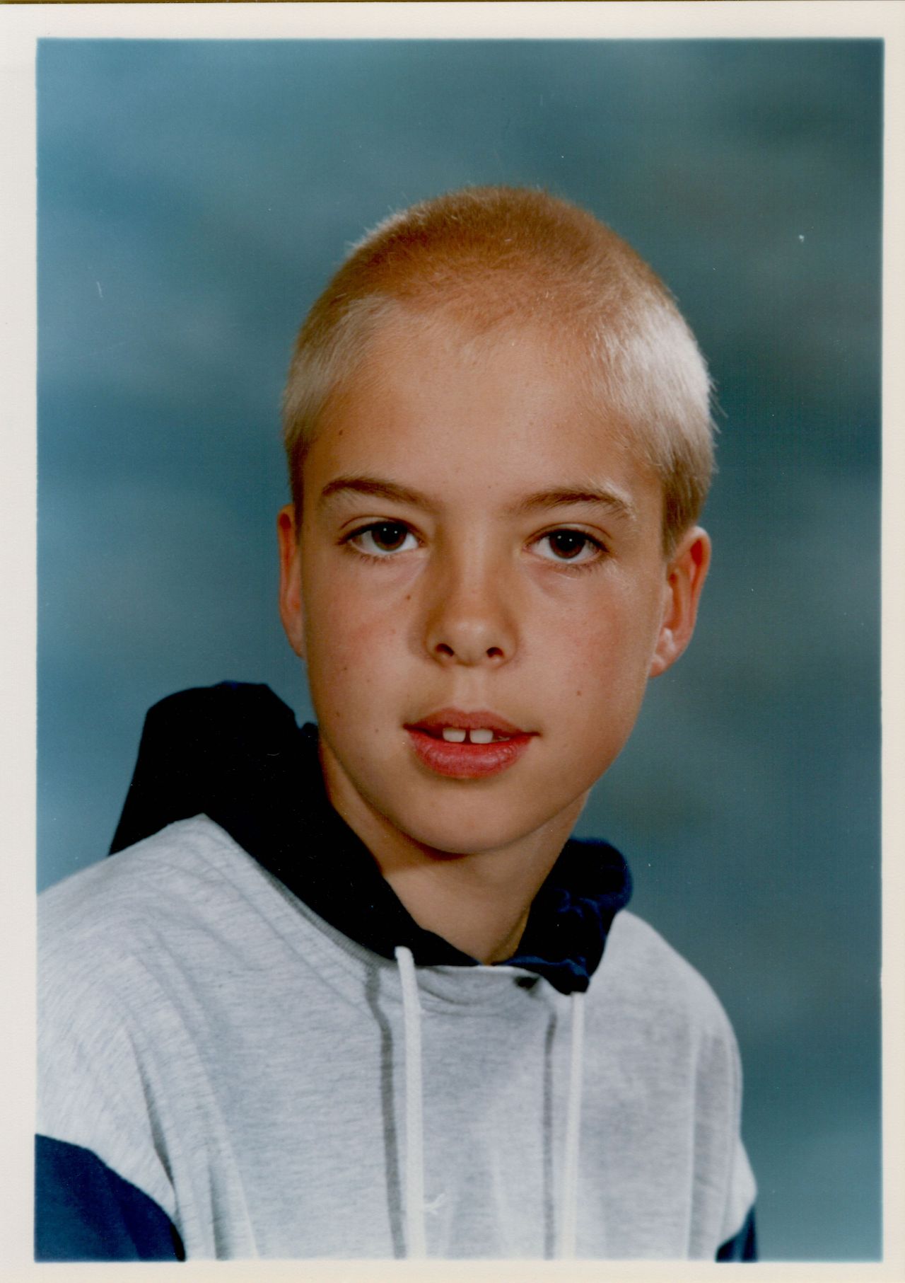 A young boy with short blond hair wears a hoodie and looks directly at the camera with a neutral expression.