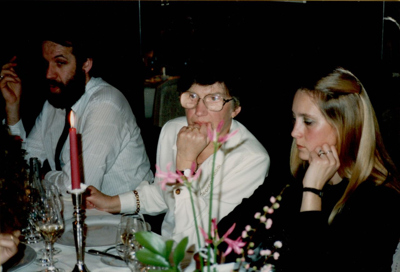 Three people sit at a dinner table with wine glasses and a lit candle, appearing engaged in conversation or thought.