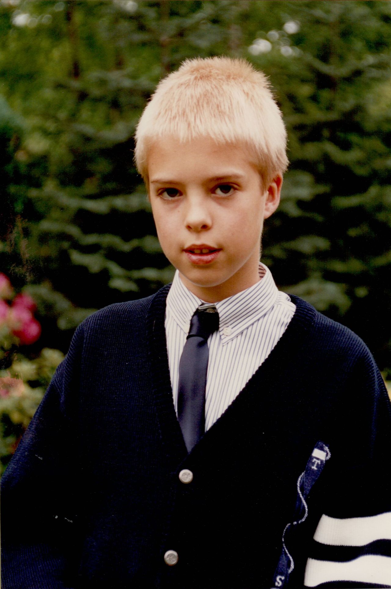 A young boy in formal attire stands outdoors, wearing a striped shirt, dark tie, and navy cardigan.
