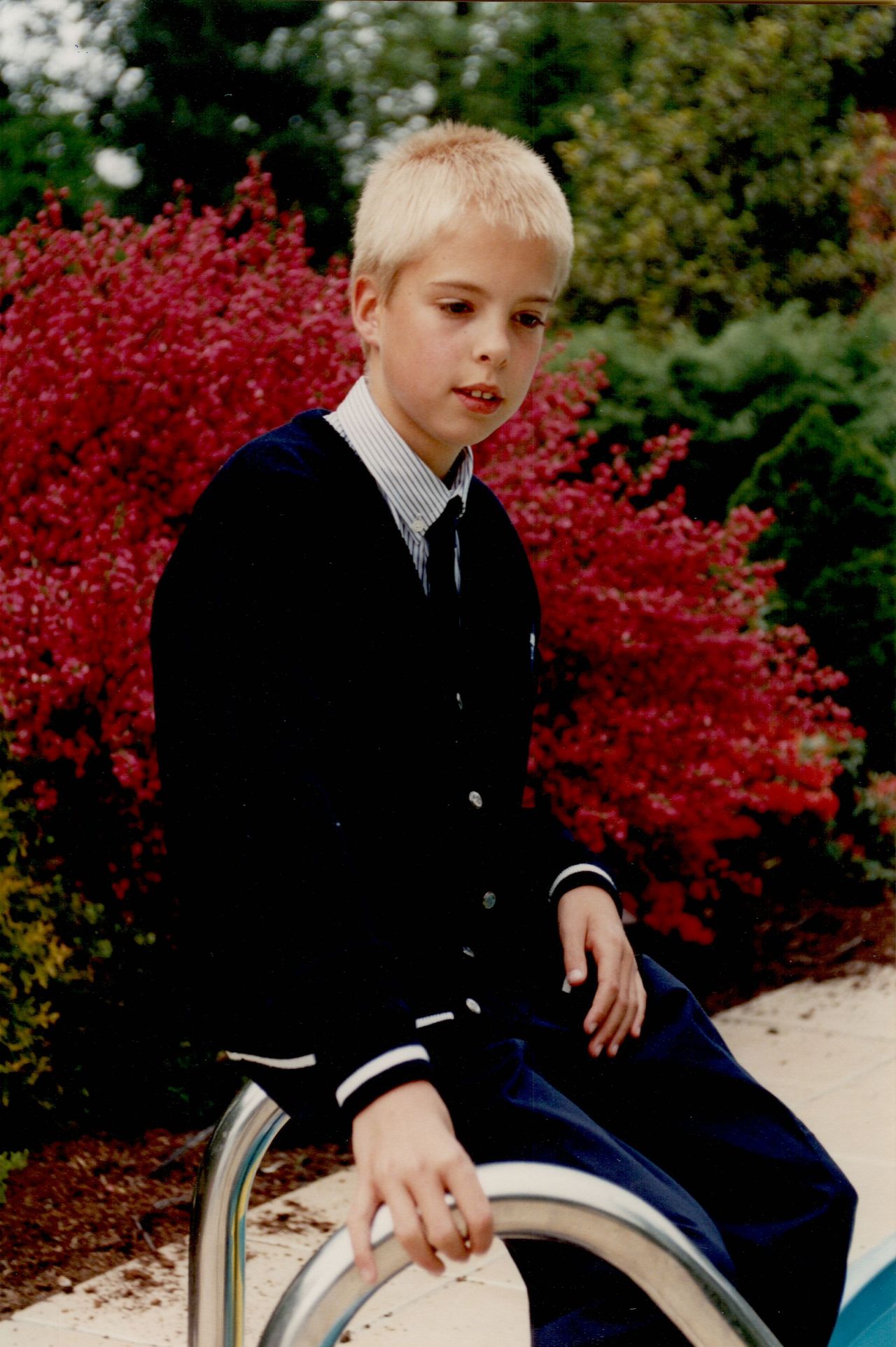 A young boy in formal clothing sits on a metal railing near a pool, looking slightly away from the camera.