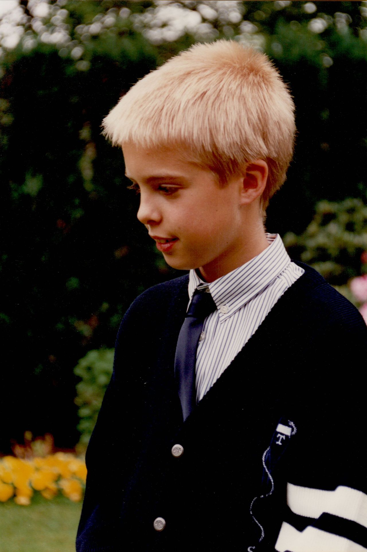 A young boy in formal attire stands outside, wearing a striped shirt, dark tie, and cardigan, looking to the side.