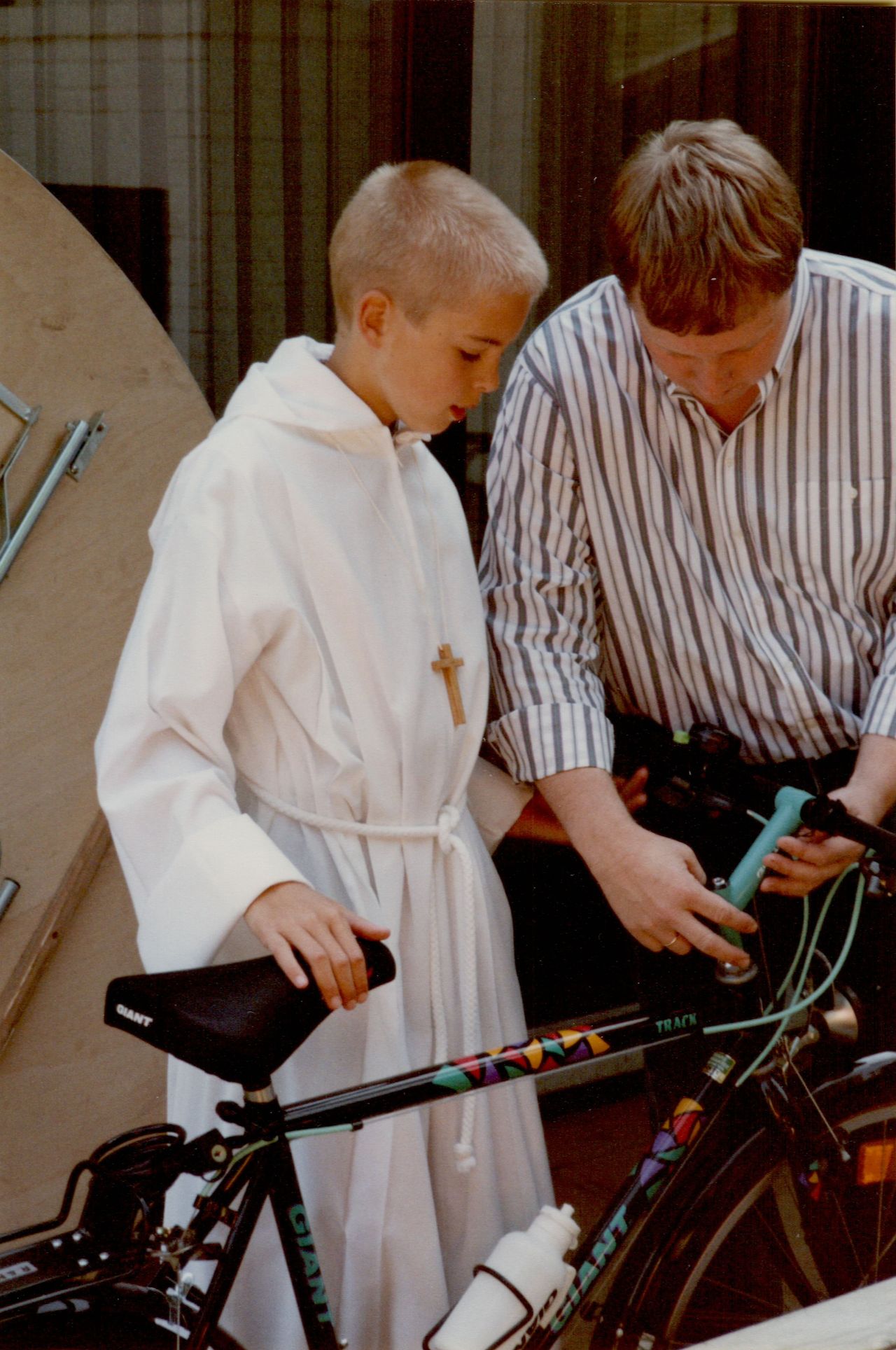 A boy in a white robe with a cross necklace stands next to a man adjusting a bicycle's handlebars.