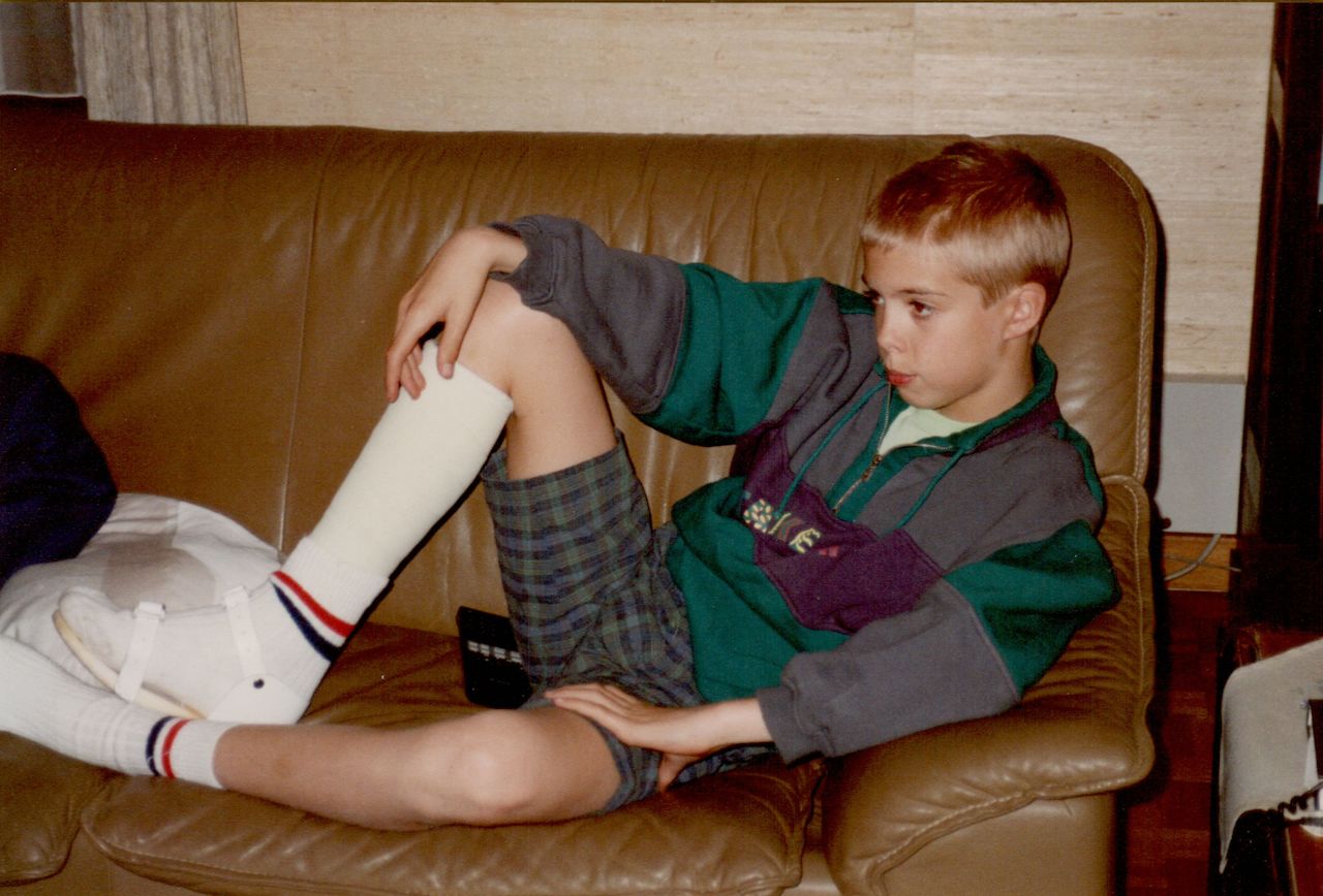 A boy sits on a couch with his leg in a cast, wearing a brace and looking to the side.