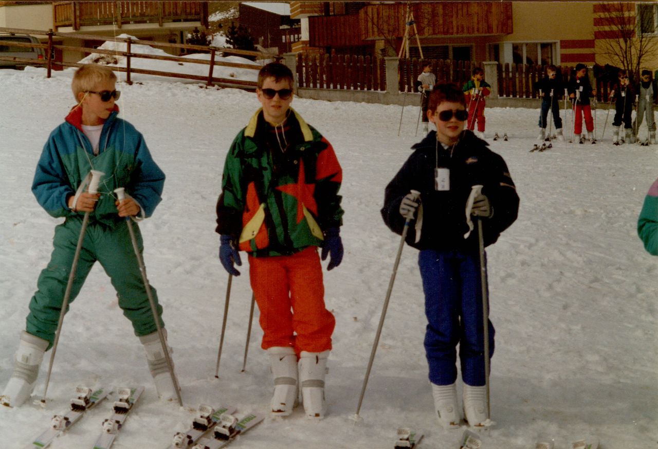 Three children in ski gear stand on a snowy slope, holding ski poles, with a group of skiers in the background.
