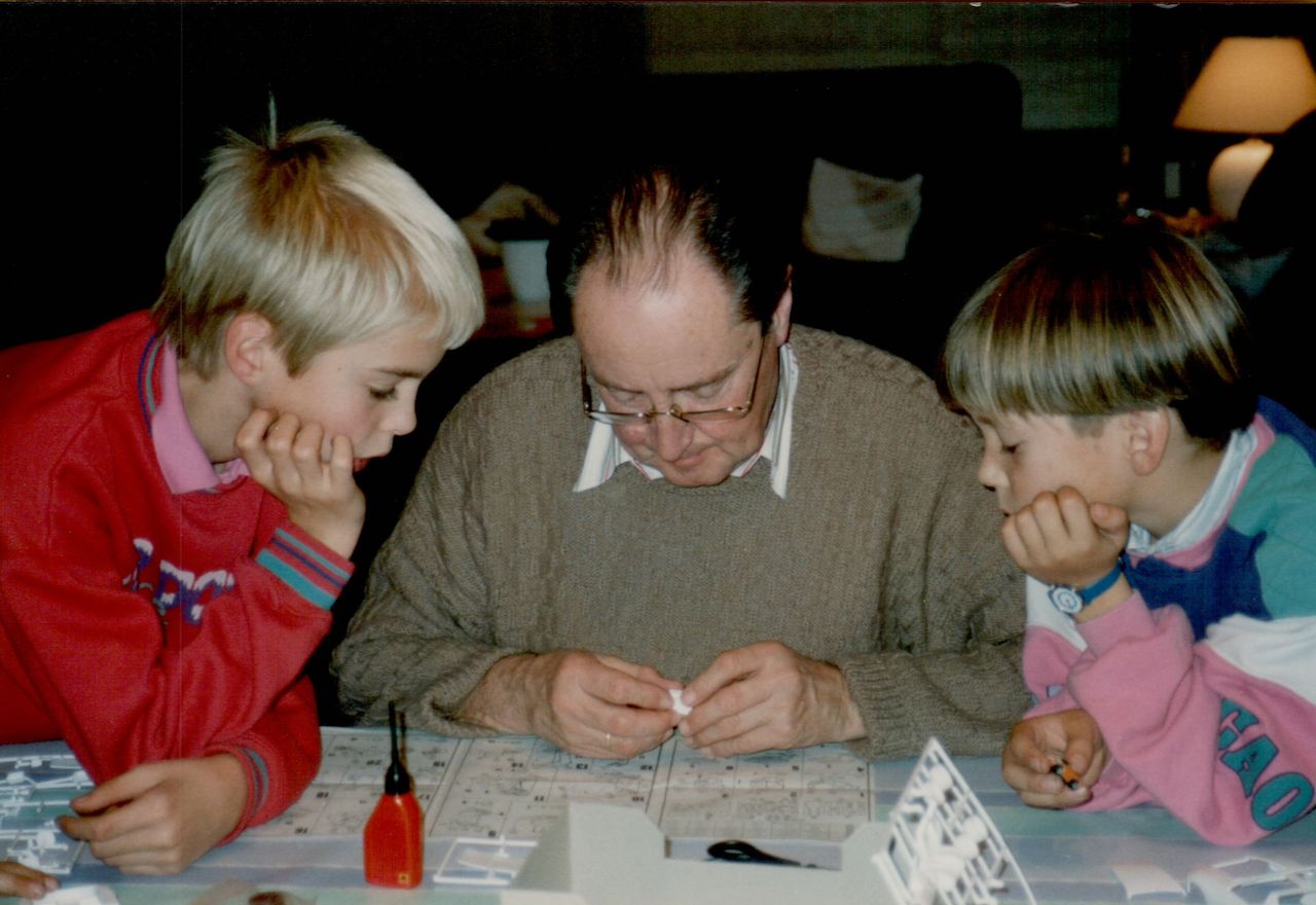An older man and two children work together on a model project, carefully assembling small pieces at a table.