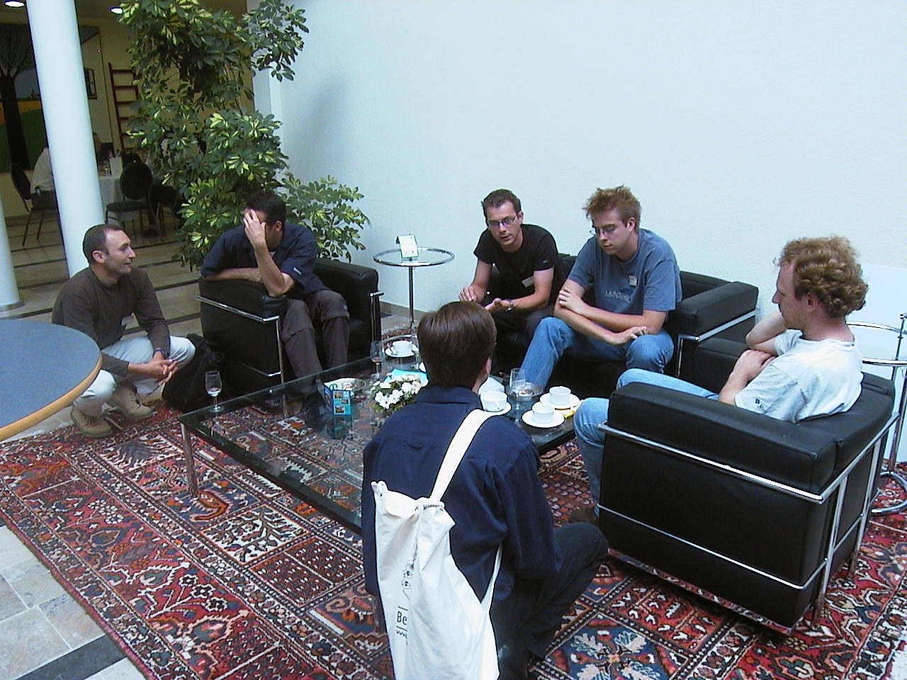 A group of six people sits in a lounge area, engaged in conversation with coffee cups on the table.