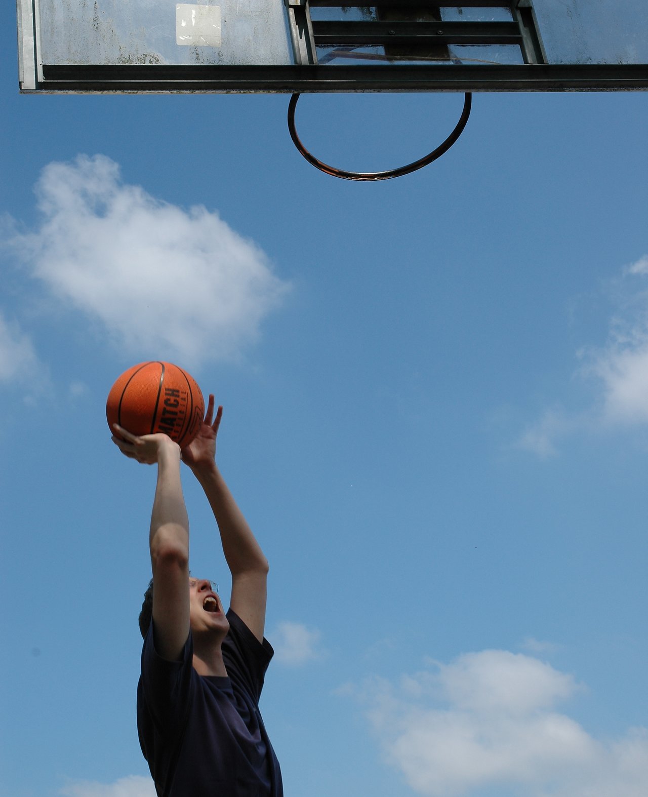 A person in a dark shirt prepares to shoot a basketball toward a hoop against a blue sky.