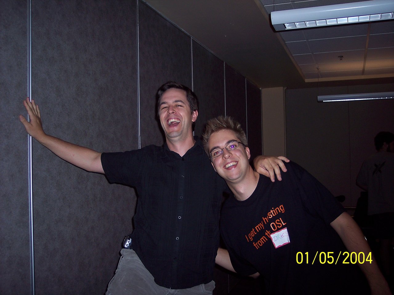 Two smiling people pose together indoors, one leaning against a wall with an arm outstretched.