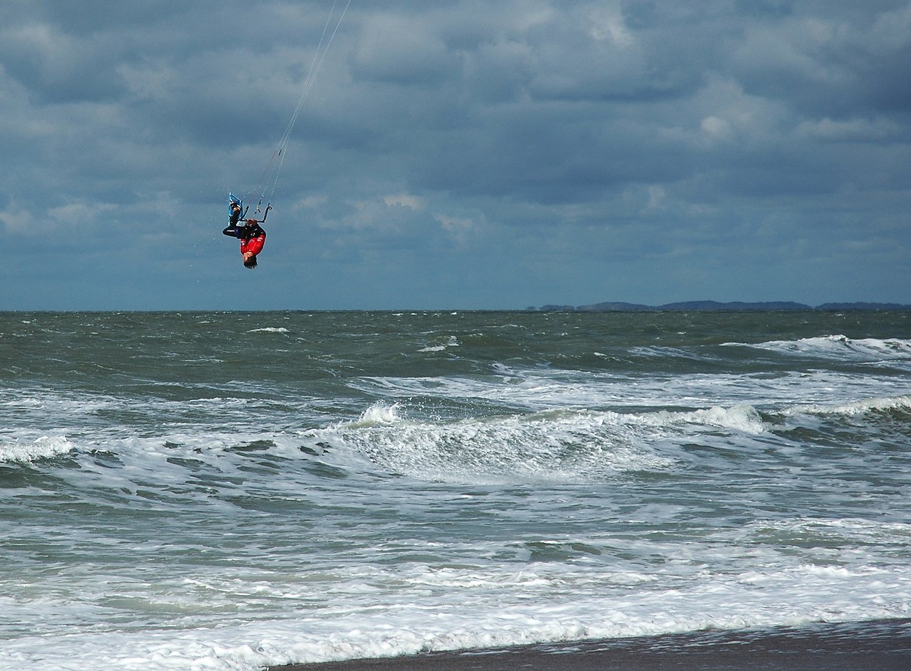 A kite surfer in a red jacket performs an aerial flip above the ocean waves on a cloudy day.
