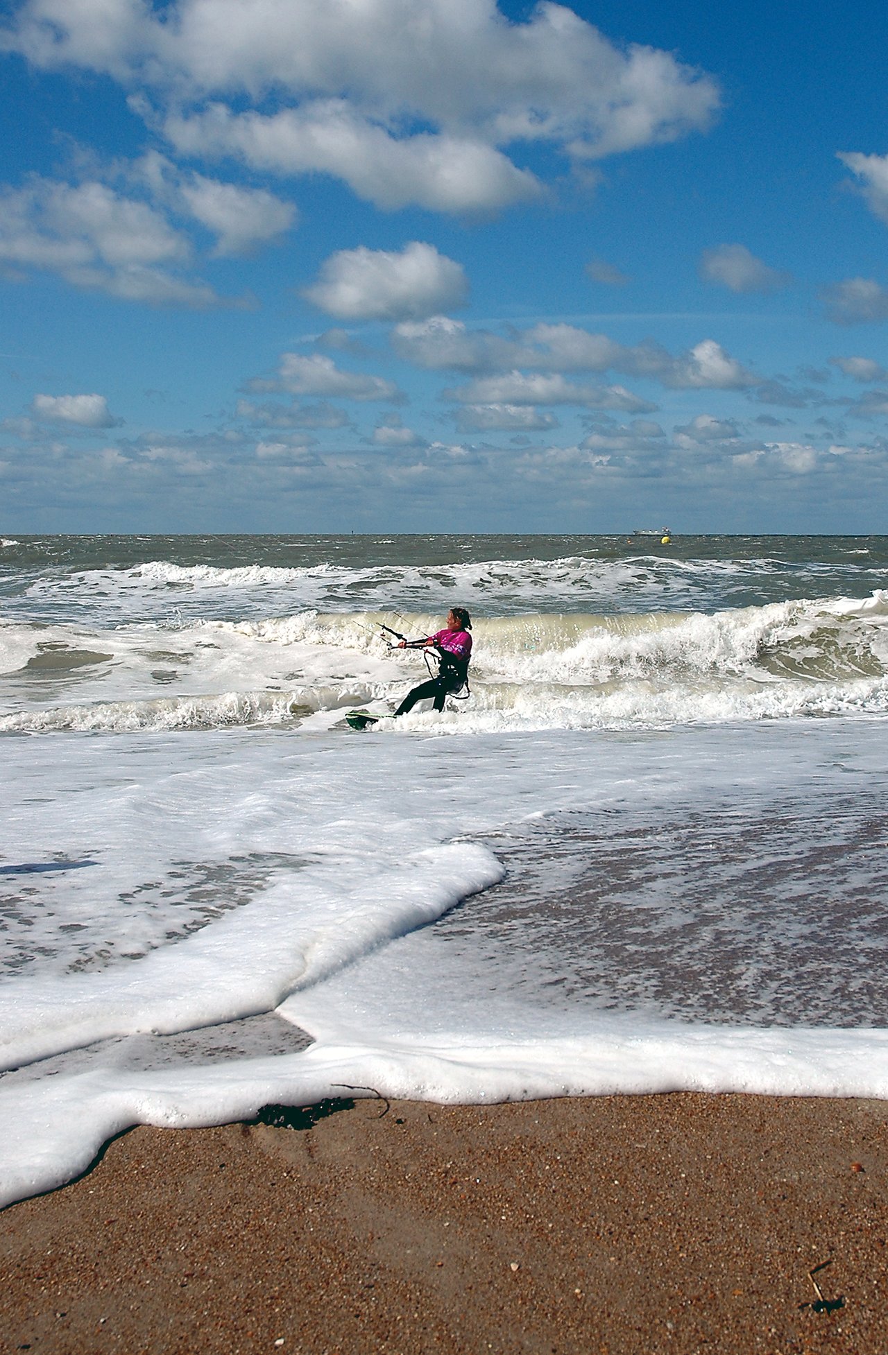 A person in a wetsuit kiteboarding on ocean waves near the shore under a partly cloudy sky.