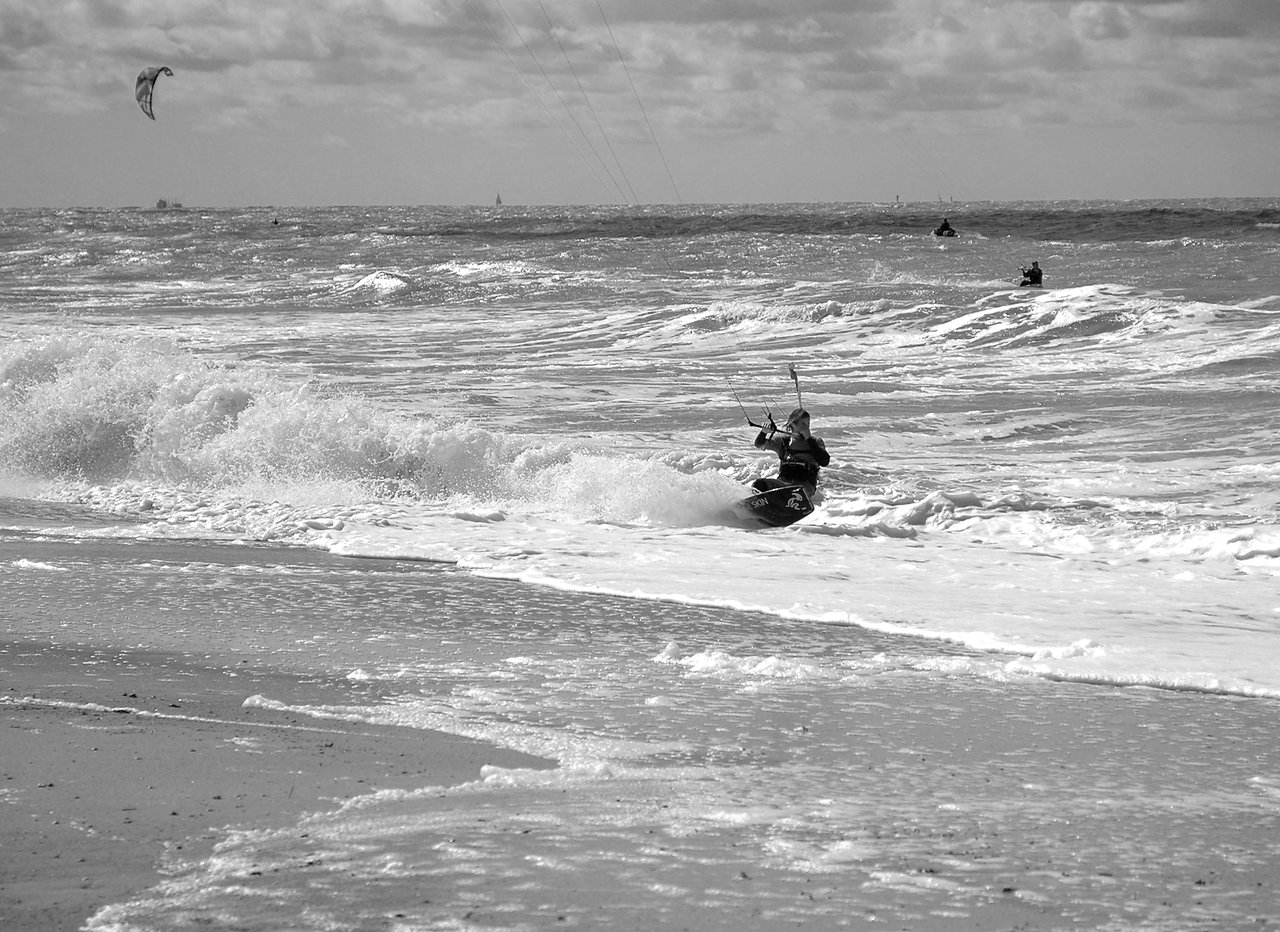 A person kiteboarding in the ocean, holding control bars while riding through waves near the shore.